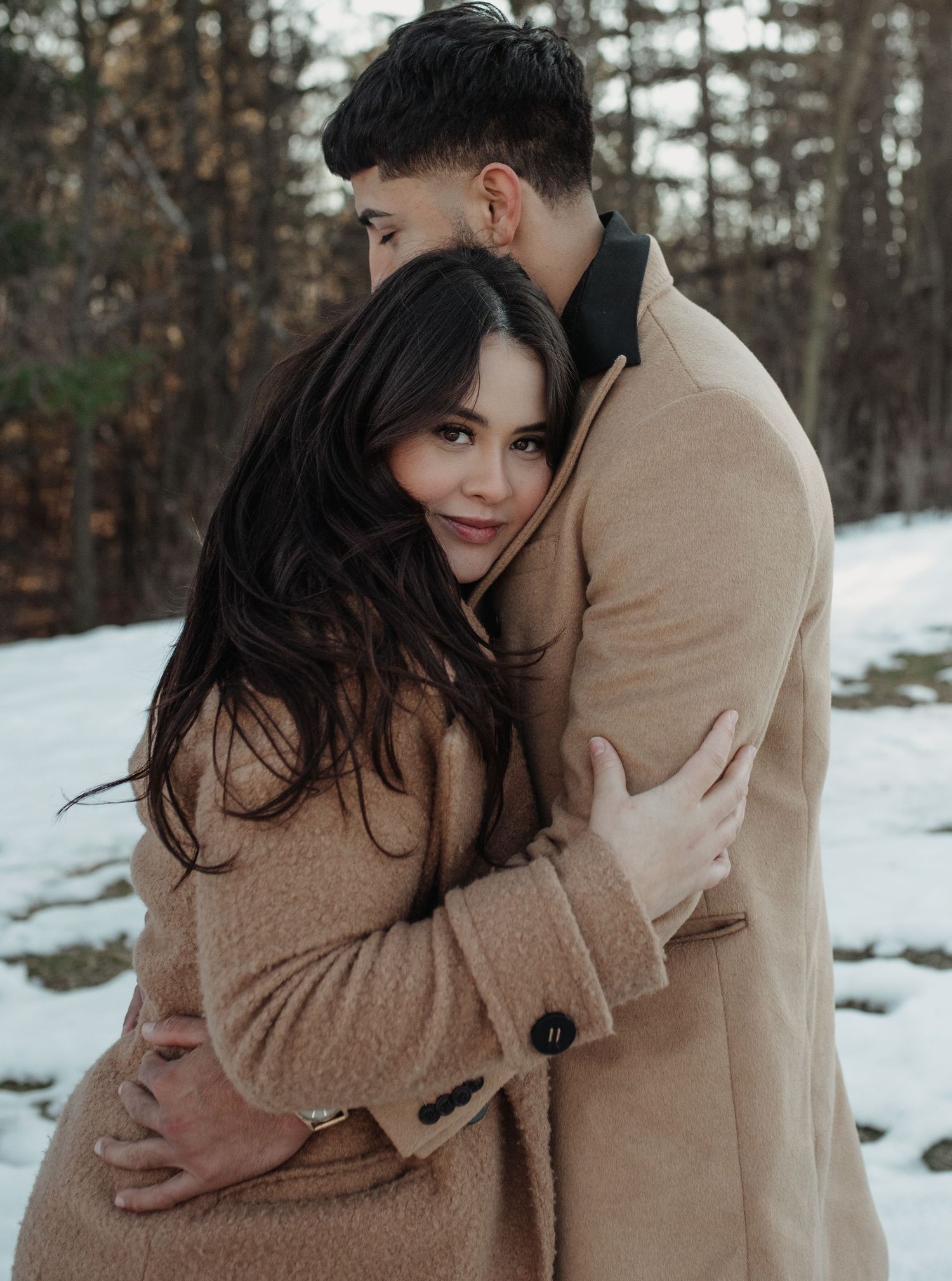 A couple in matching tan coats share affectionate moments against a snowy winter forest backdrop. Appleton, Green Bay & Kaukauna