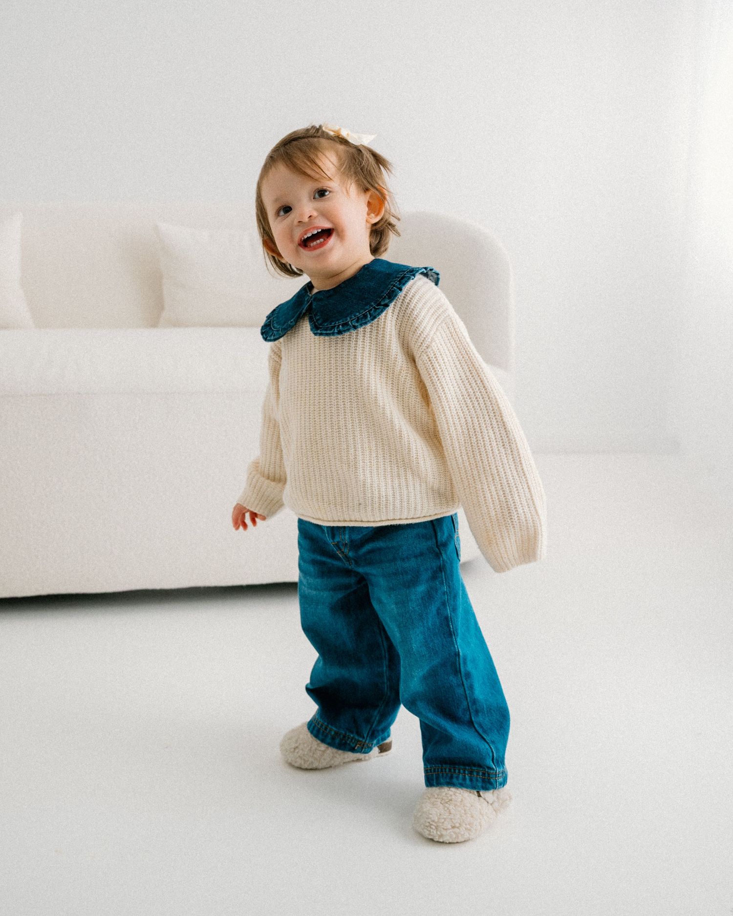 Baby in cream sweater and blue pants playing on white furniture. Appleton, Green Bay & Kaukauna