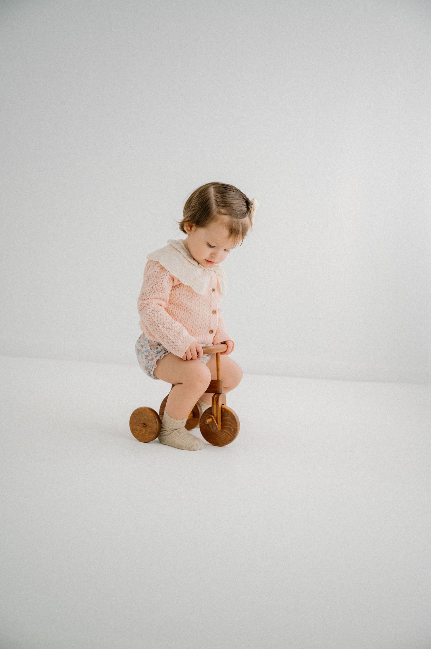 Child wearing pink cardigan on wooden tricycle against white background. Appleton, Green Bay & Kaukauna