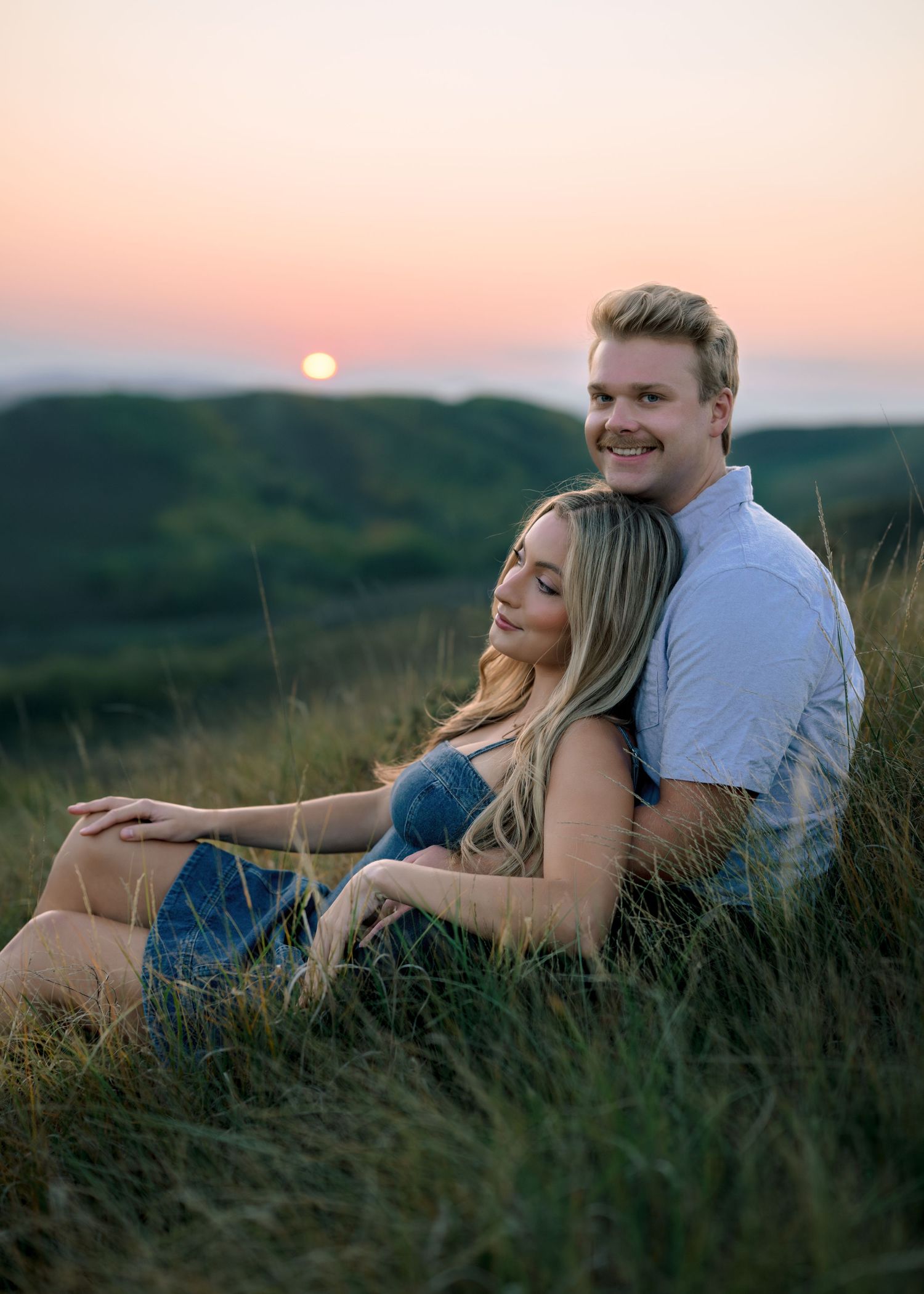 A couple lying together in tall grass during a romantic sunset photoshoot with pink sky in the background.