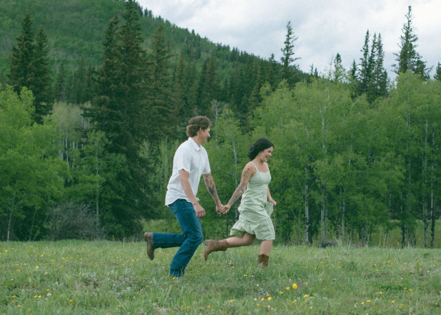 A couple running playfully through a green meadow with mountains and evergreen trees in the background. Calgary Alberta Engagement Photo