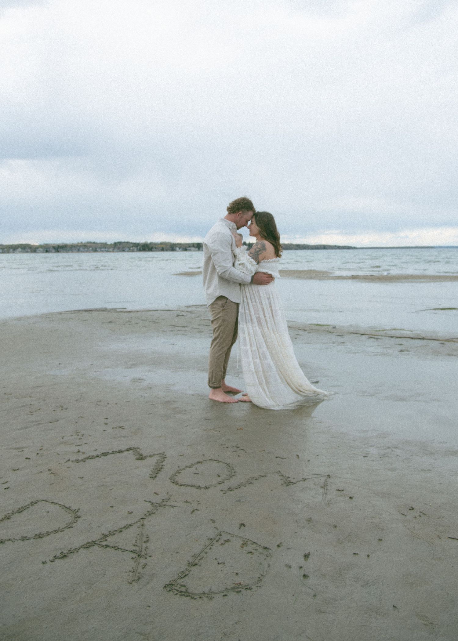 A couple in white attire shares a romantic moment on a sandy beach with the ocean behind them. Calgary Alberta Maternity Photographer.