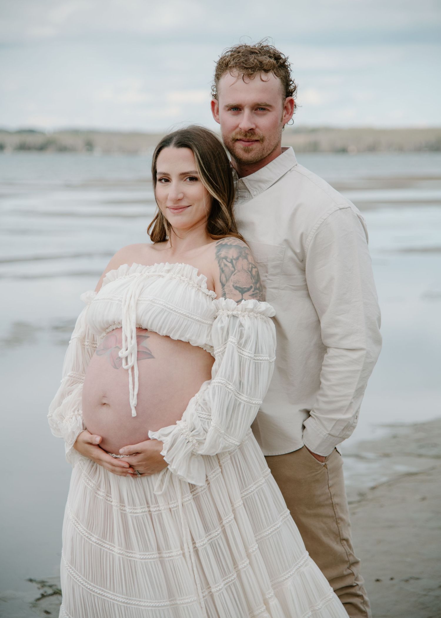 Expectant couple poses for a maternity photo on a beach, wearing white and beige clothing. Calgary Alberta Maternity Photographer.