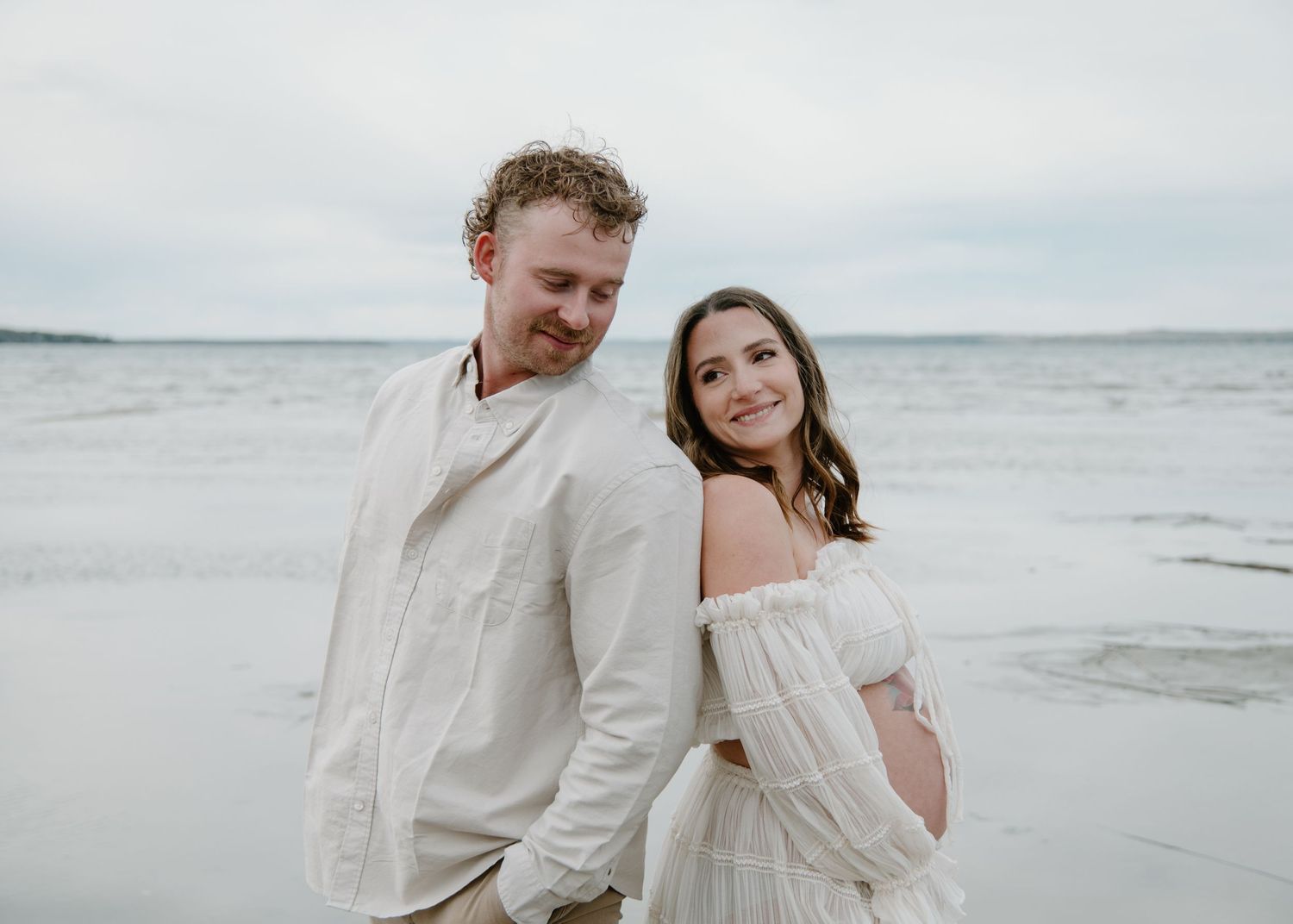 A beach maternity photoshoot capturing a joyful moment between a couple in matching light coloured outfits. Calgary Maternity Photographer