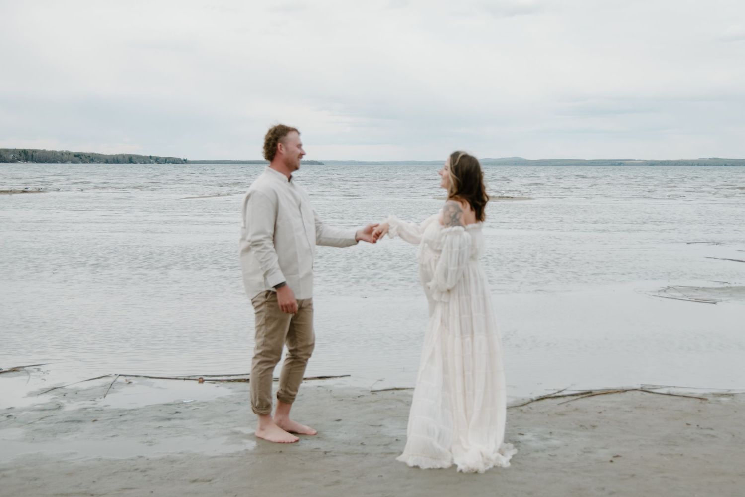 A couple holding hands while standing on a beach with waves and cloudy skies in the background. Calgary Alberta Maternity Photographer.