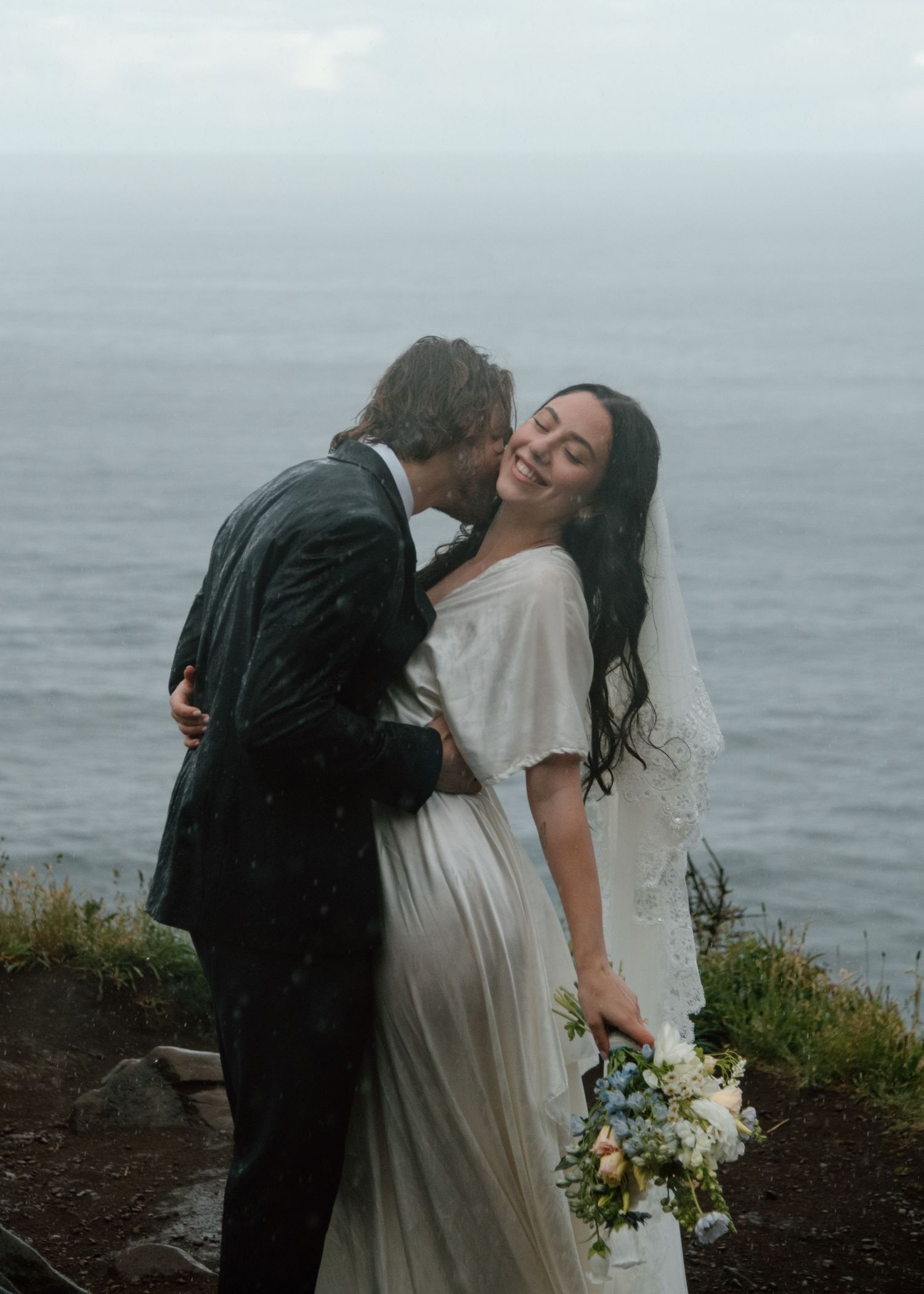 Wedding couple poses dramatically on cliff edge overlooking misty ocean. Calgary Wedding Photographer