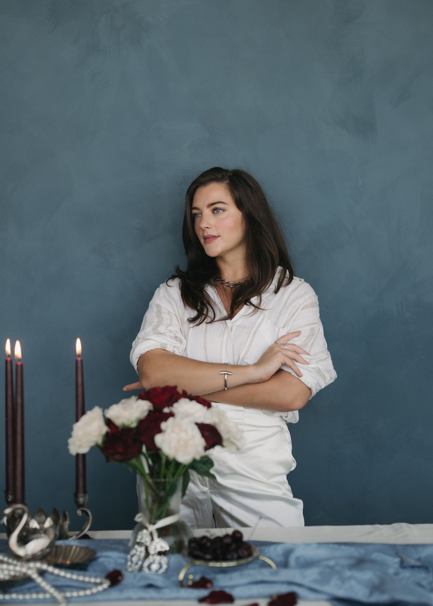 Woman in white shirt sits at moody table setting with dark candles and flowers against blue-gray wall.