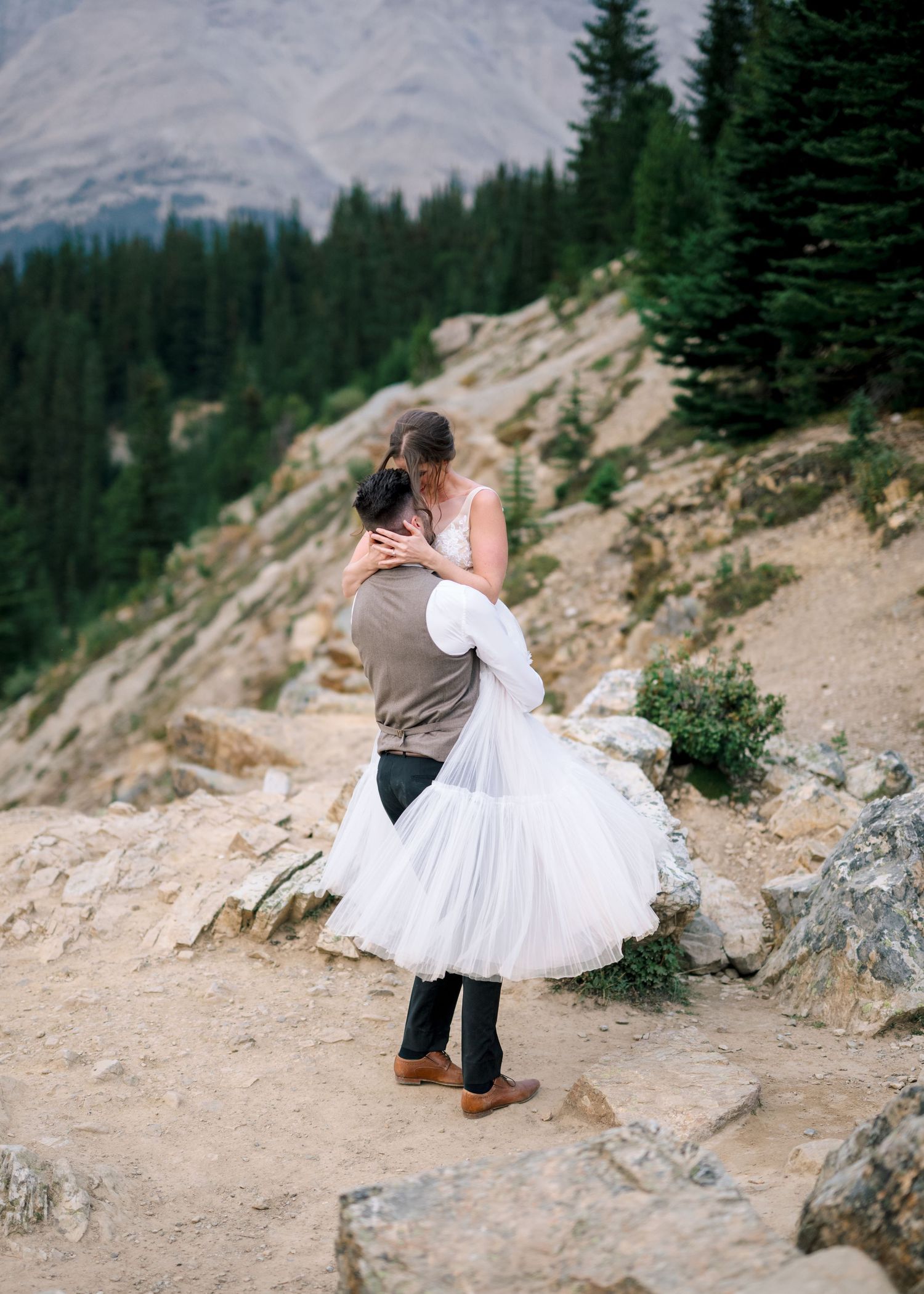 A couple shares an intimate dance moment on a scenic mountain path surrounded by evergreen trees and rocky terrain.