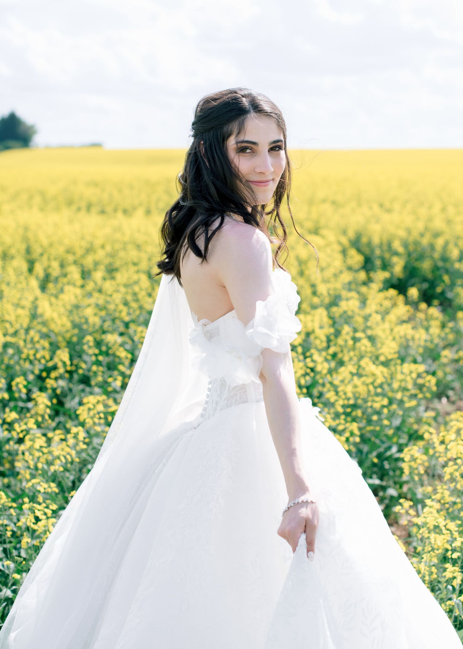 A person in a flowing white dress poses gracefully in a vibrant yellow canola field on a sunny day.