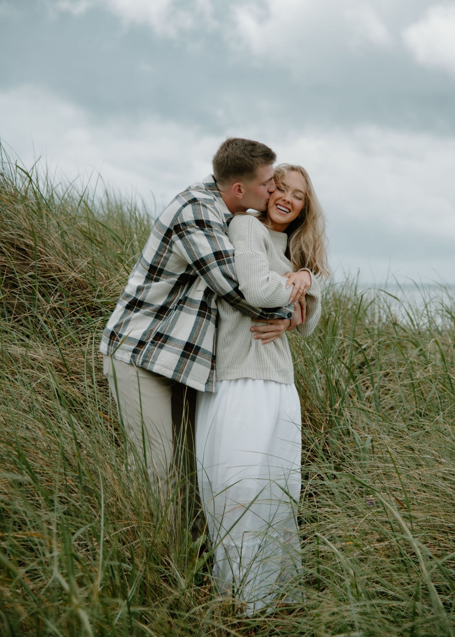 Loving couple dances together in field of tall grass beneath overcast sky on Oregon Coast. Calgary Lifestyle and Engagement Photographer.