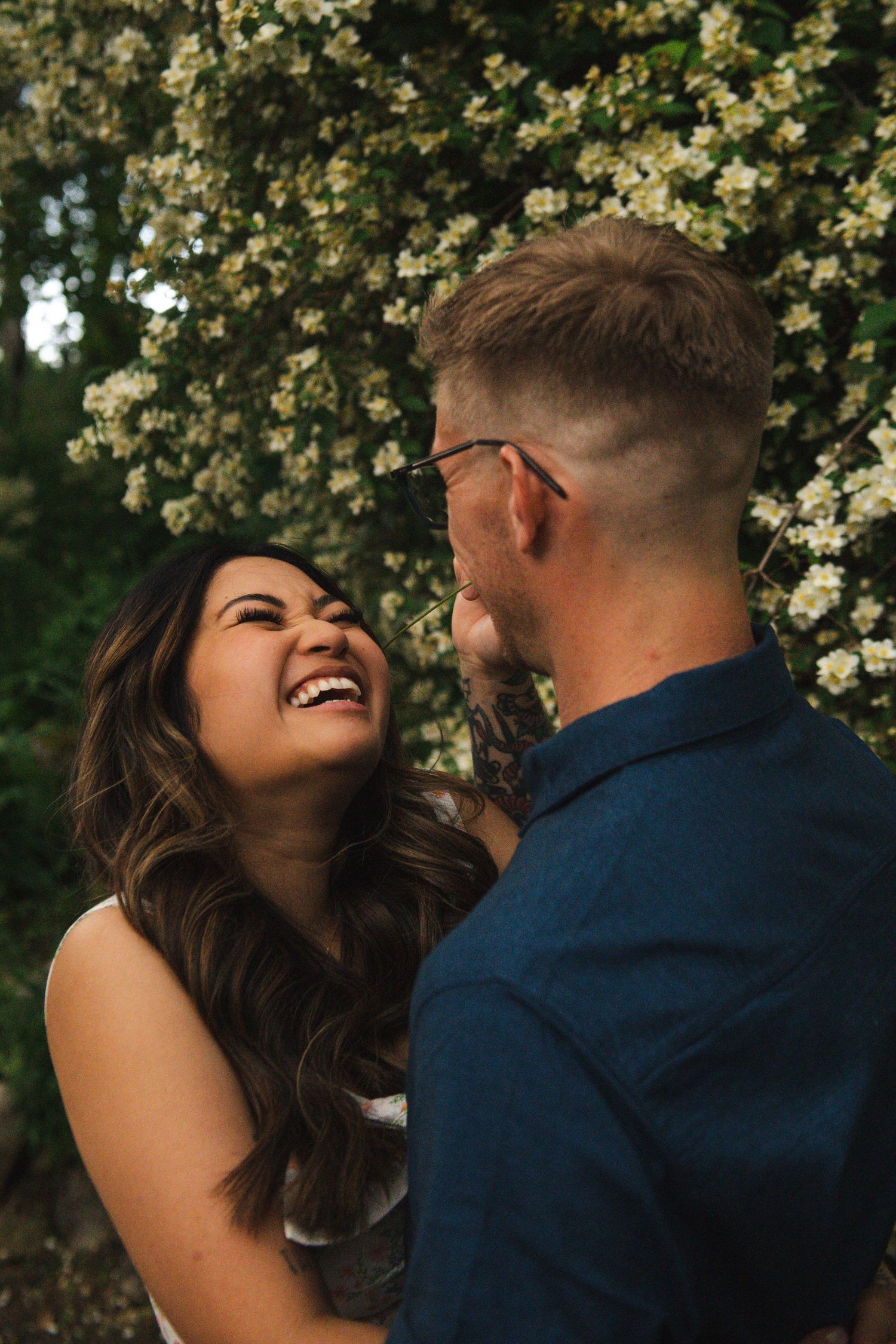 A couple shares joyful laughter under blooming spring tree branches in Calgary. Calgary Lifestyle and Engagement Photographer.