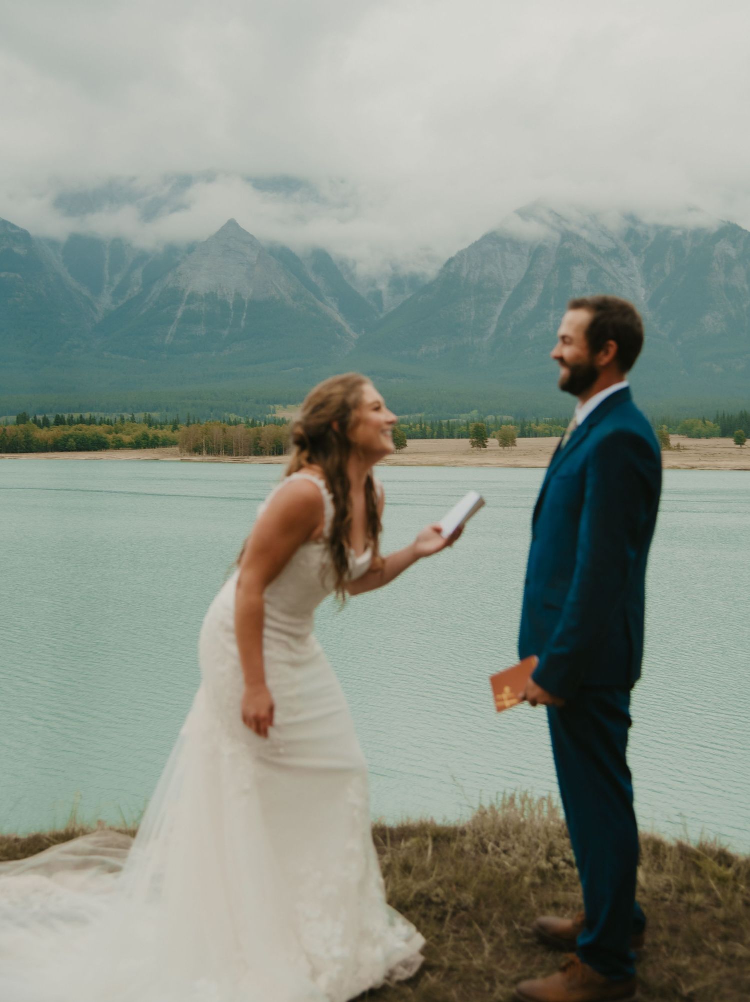 Wedding couple shares a candid moment by a mountain lake with snow-capped peaks in the distance. Calgary Wedding Photographer