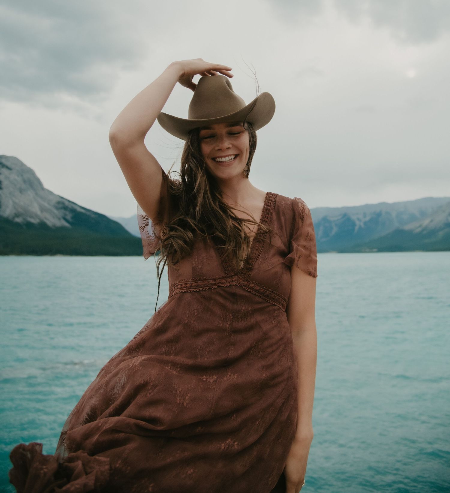 Woman in brown dress posing by a turquoise lake with mountain views. Calgary Branding Photographer