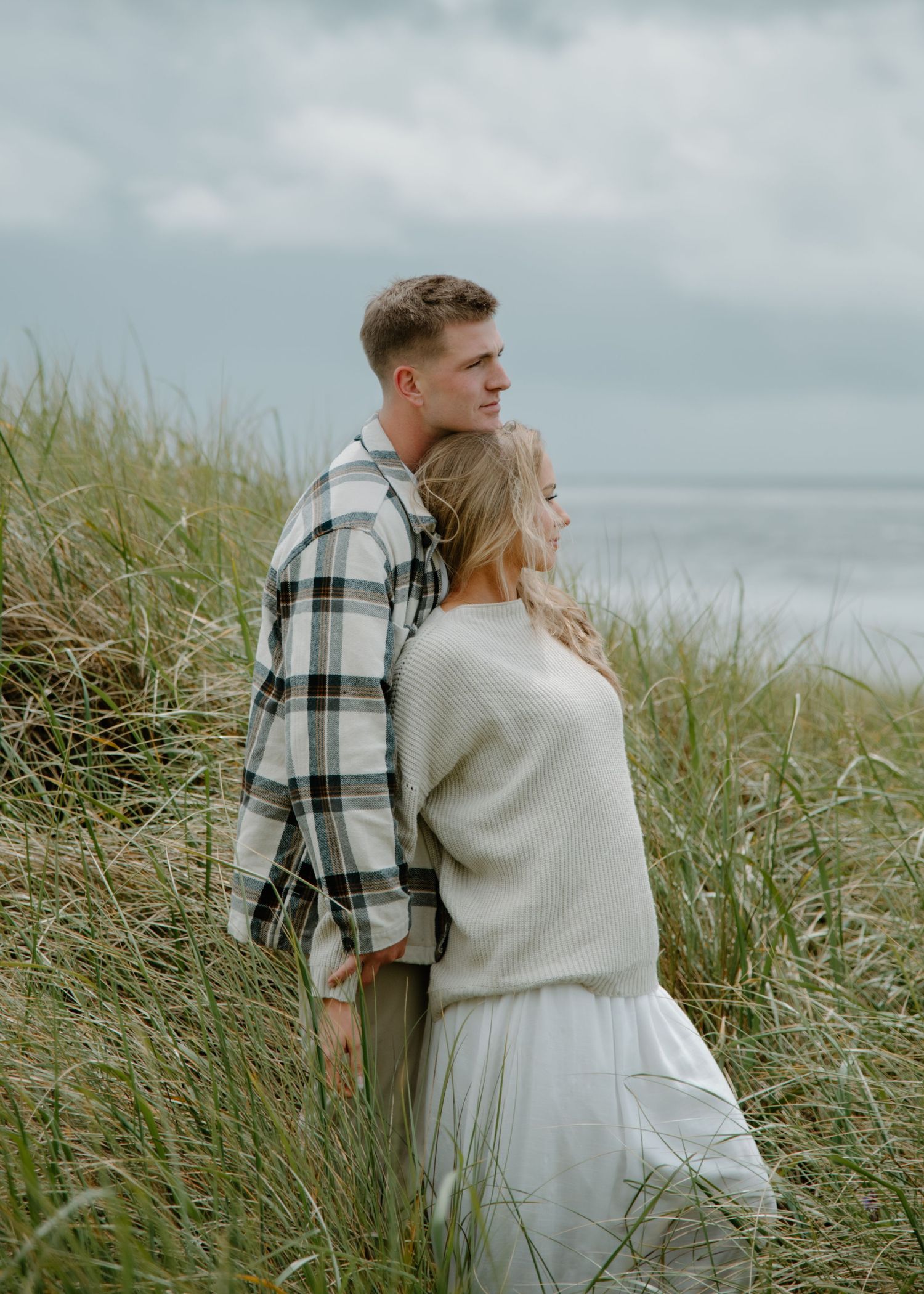 Tender moment captured between two people on windy coastal dunes on the Oregon Coast. Calgary Alberta Engagement Photographer.