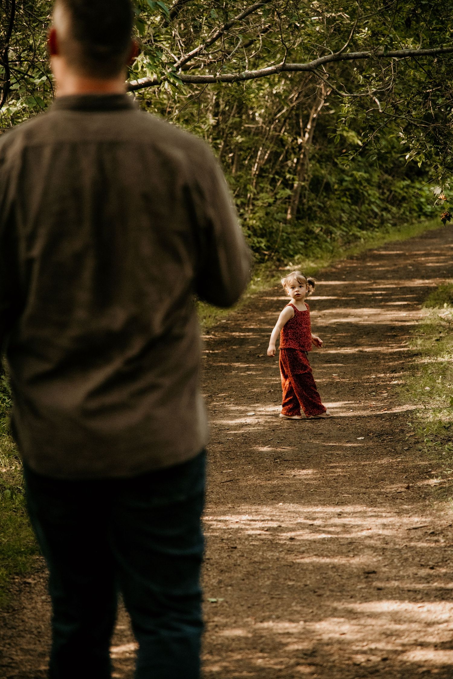 Child in red outfit walks away on a sunlit dirt path through wooded area. Calgary Alberta Family Photographer.