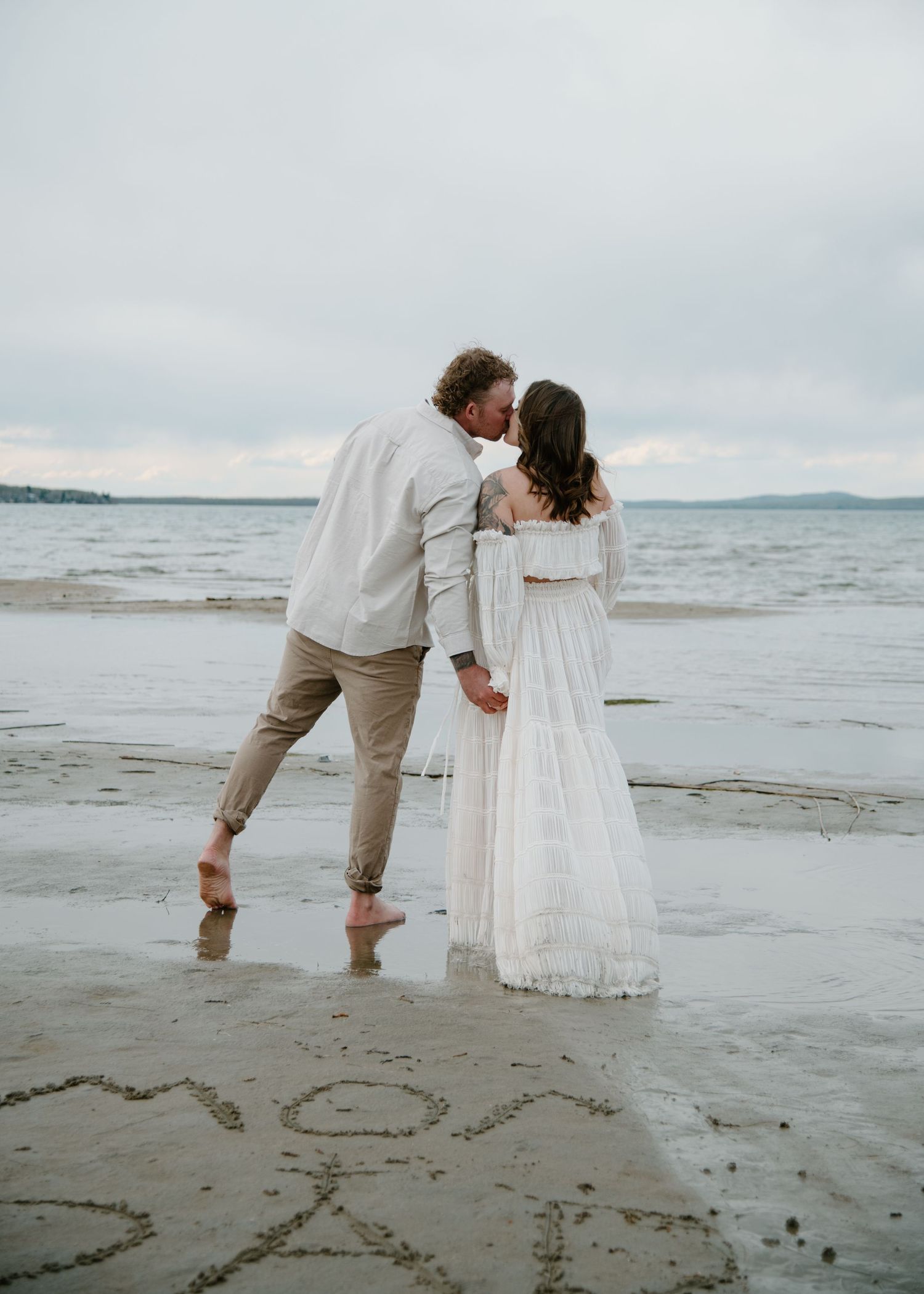A couple in white standing on wet beach sand with writing visible below. Calgary Alberta Maternity Photographer.