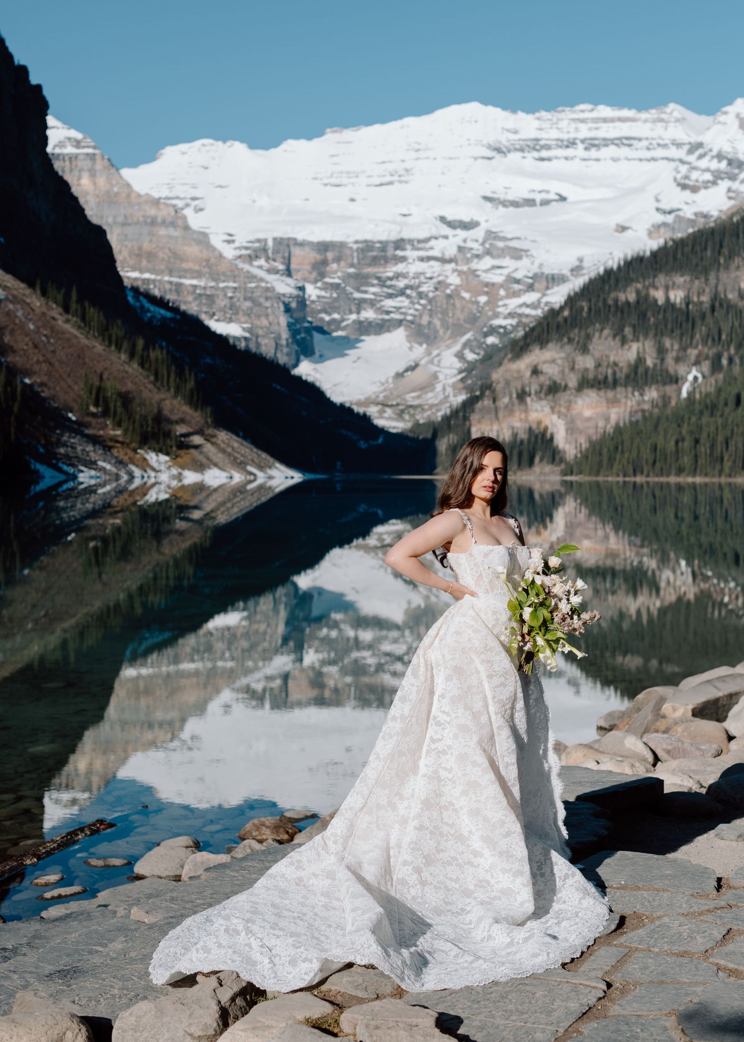 Bride in lace gown poses at Lake Louise with snow-capped mountains reflecting in turquoise water. Banff and Calgary Wedding Photographer