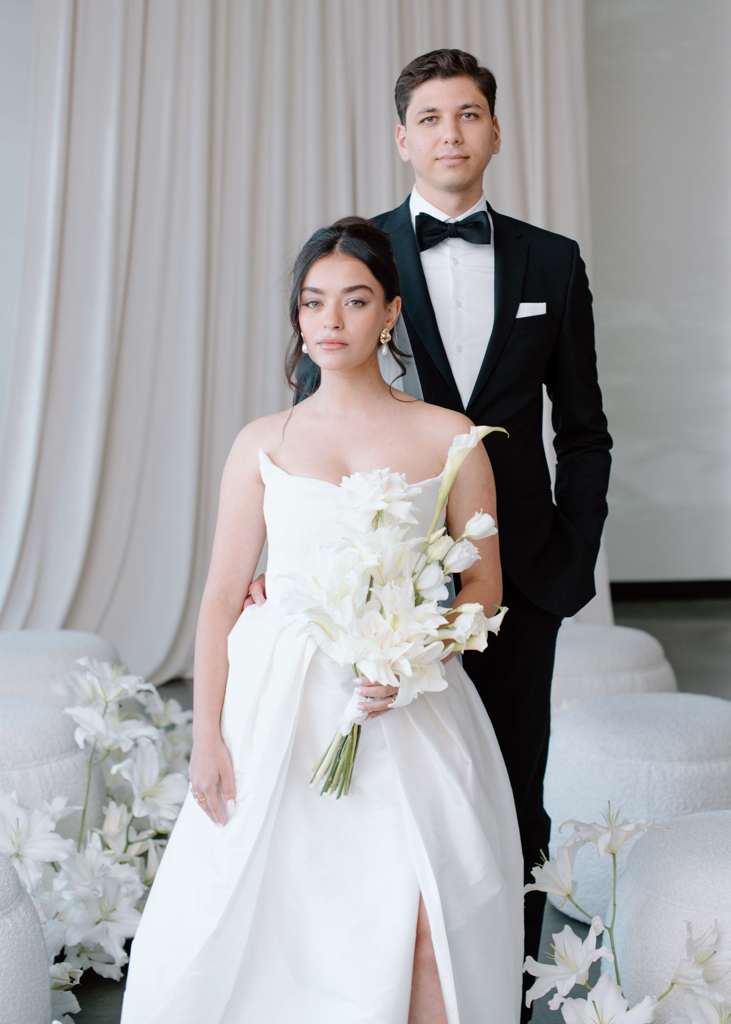 A couple in elegant wedding attire pose together with white flowers against light curtains in a modern setting.
