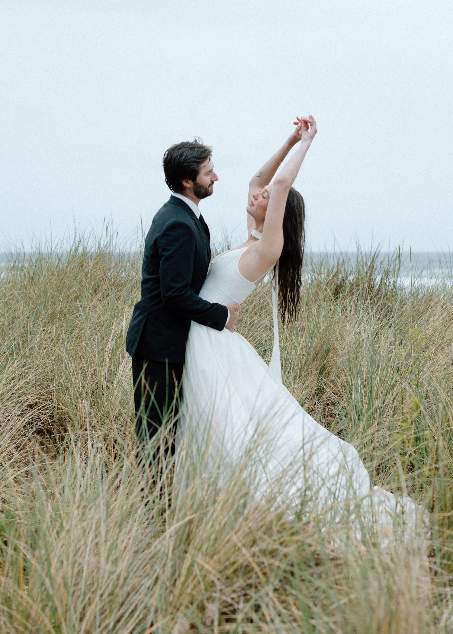 A couple dances together among tall beach grass with the ocean visible in the distance on their wedding day.