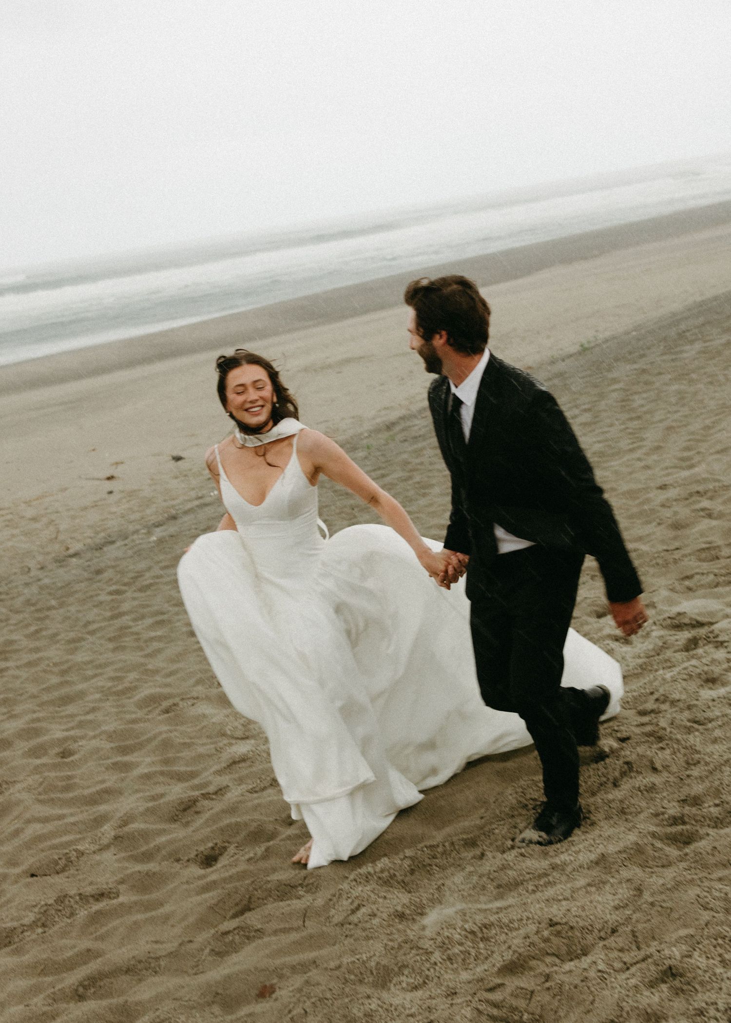 A couple in wedding attire holds hands and walks along a misty beach shoreline in sequence of romantic candid moments.