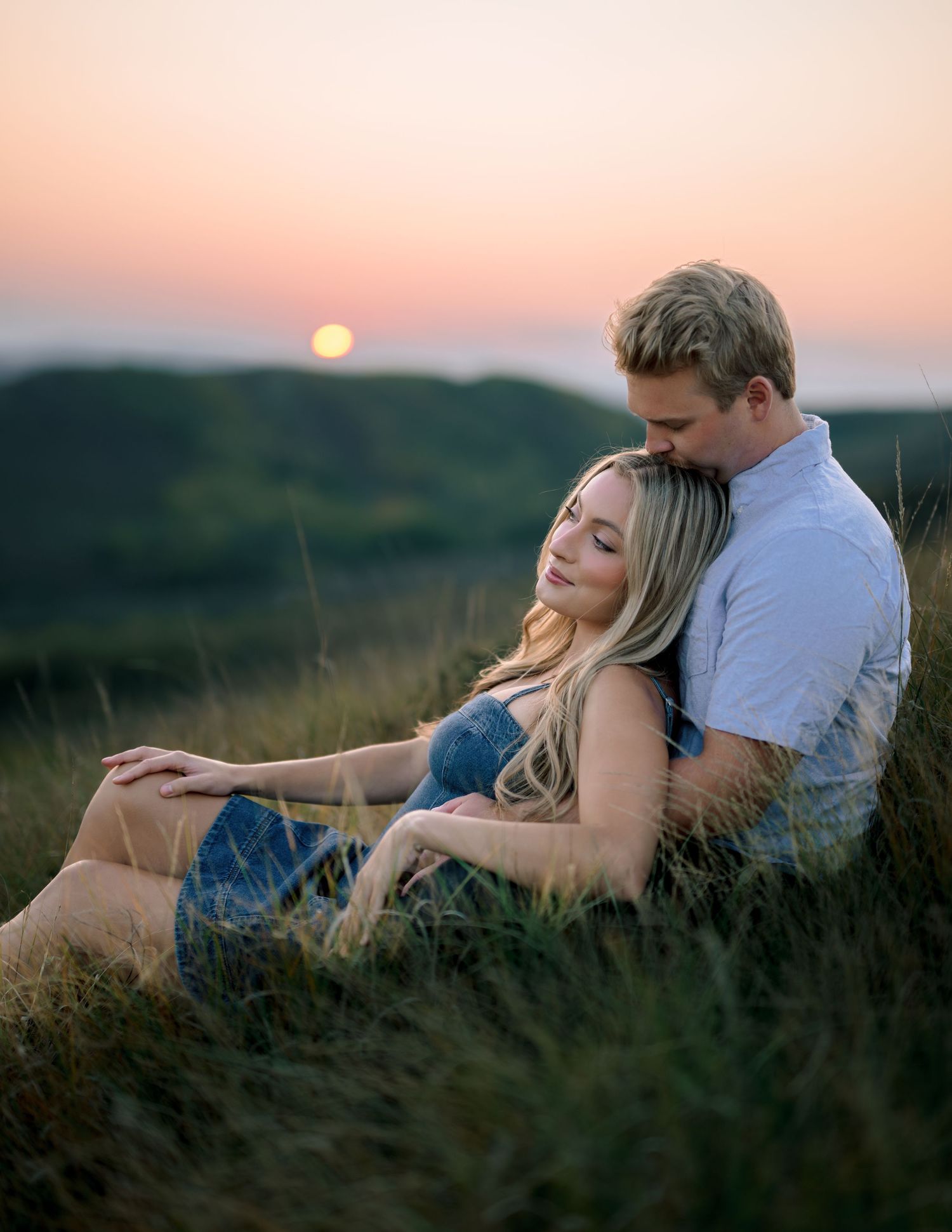 A couple embraces on a grassy hillside during sunset with a glowing orange sky in the background.
