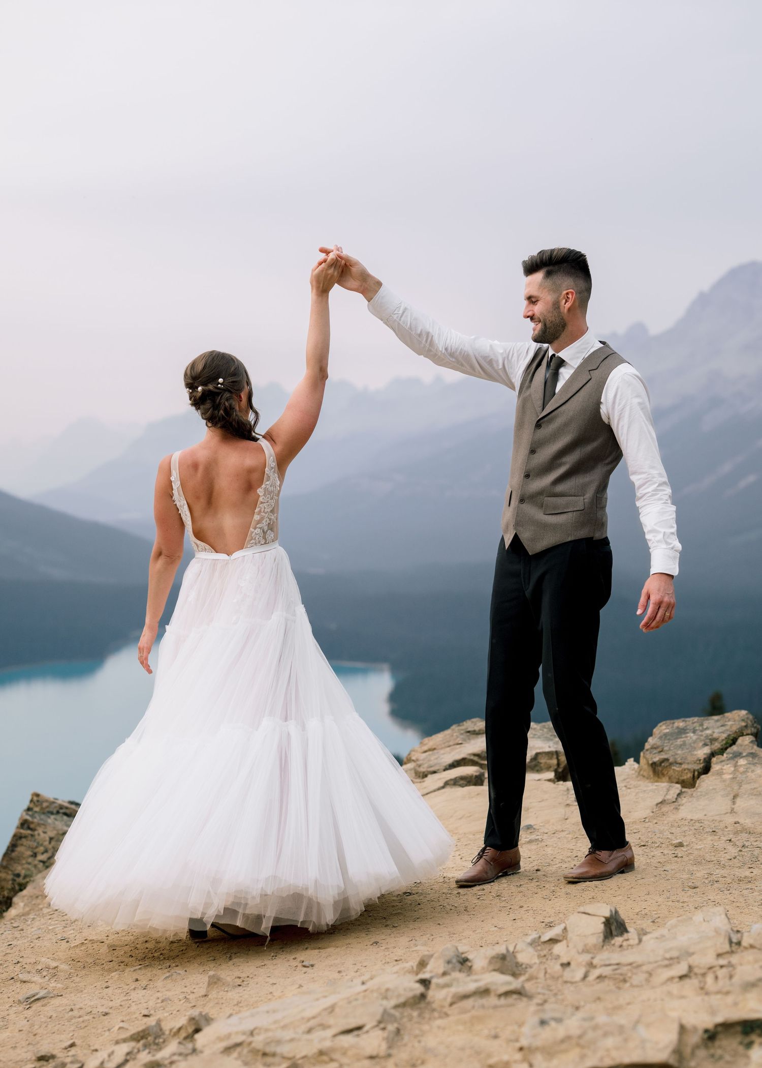 Newlyweds dance playfully on a rocky mountain ledge overlooking a misty lake and mountain landscape.