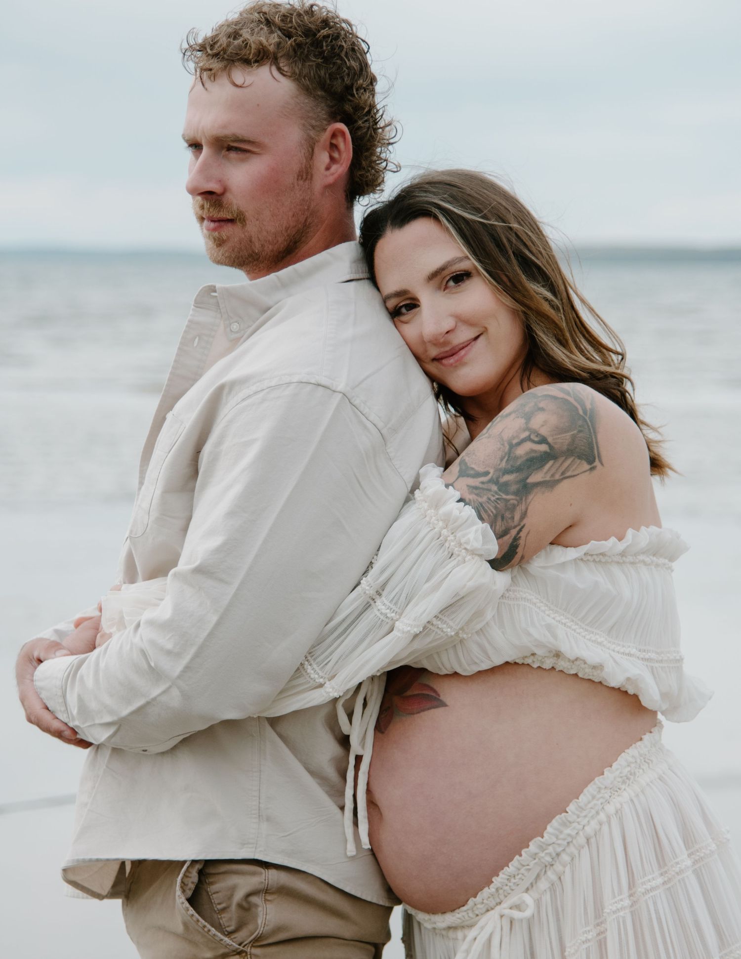 A tender moment captured between a couple in white clothing on a serene beach setting. Calgary Alberta Maternity Photographer.
