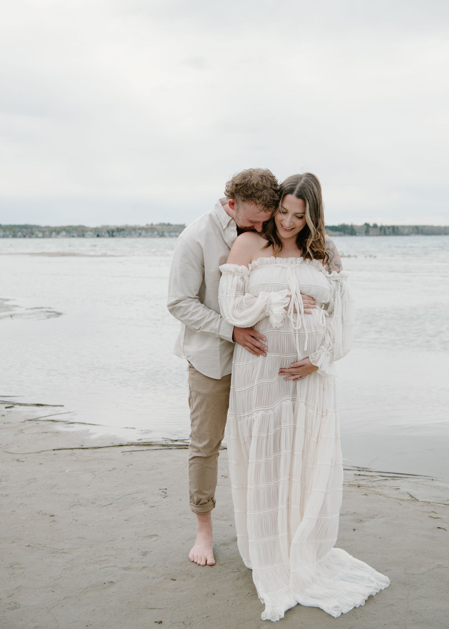 Candid maternity photos of a couple embracing on a beach with water and distant shores in view. Calgary Alberta Maternity Photographer.