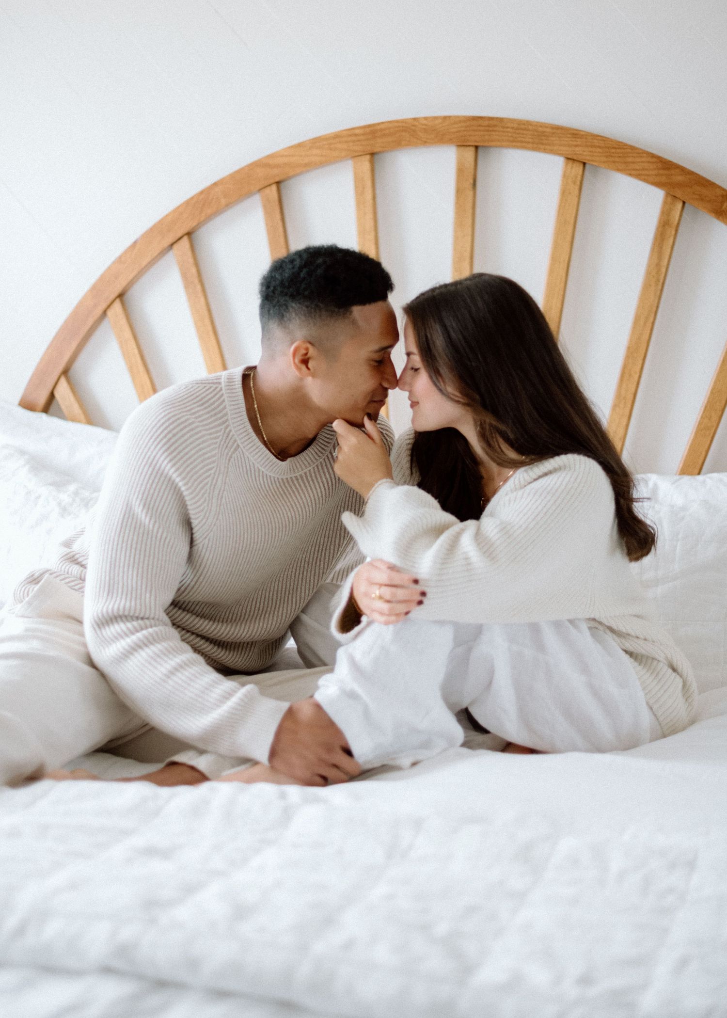 Couple in cozy white sweaters sharing an intimate moment on bed with wooden headboard.