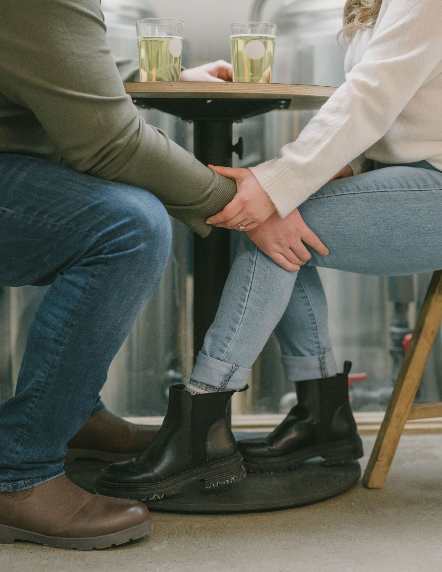 Two people sit close together at a cafe table, holding hands, wearing jeans and boots with drinks on the table above.