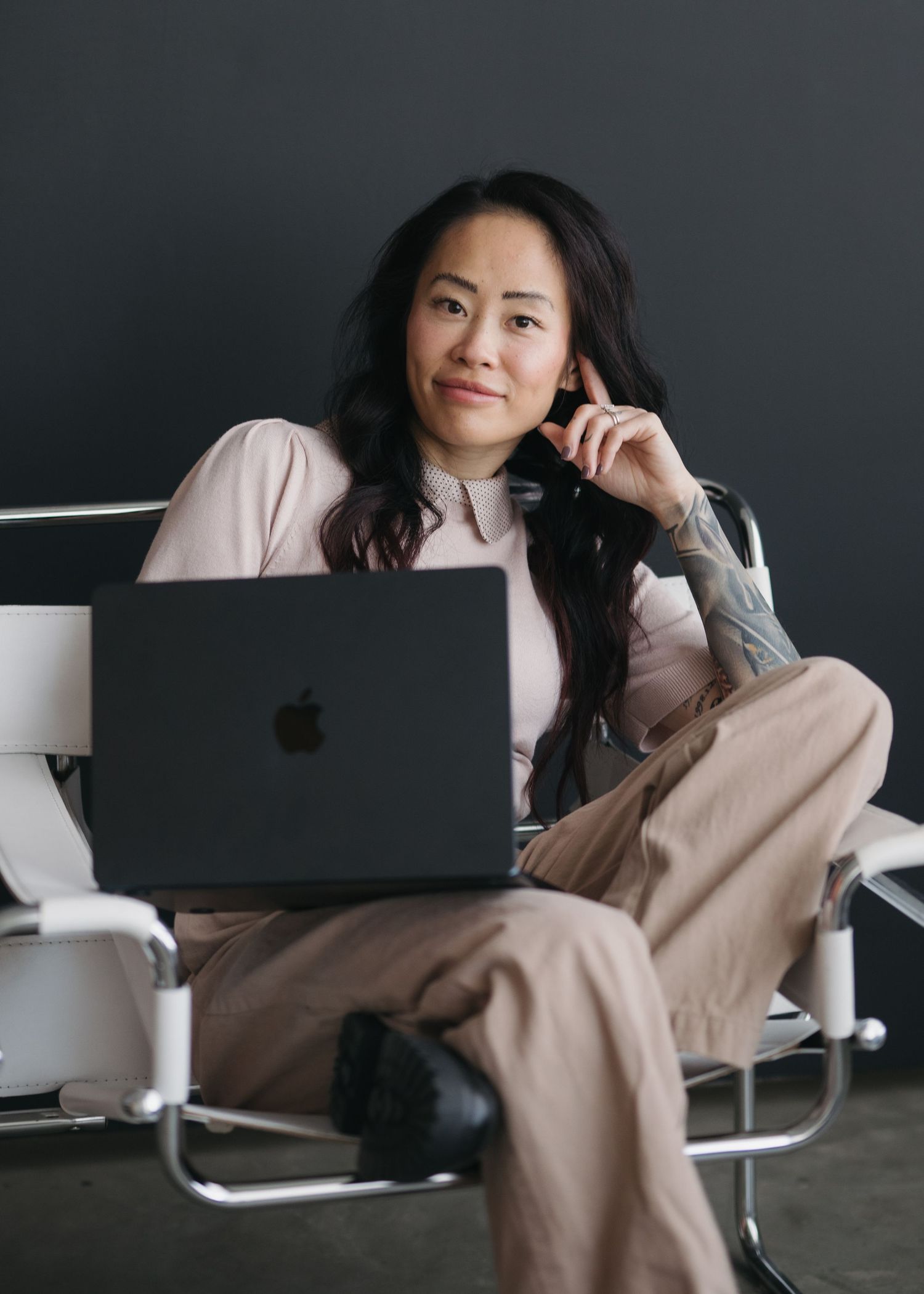 A person in a pink sweater works on a laptop while relaxing on a couch in a minimalist dark room setting.