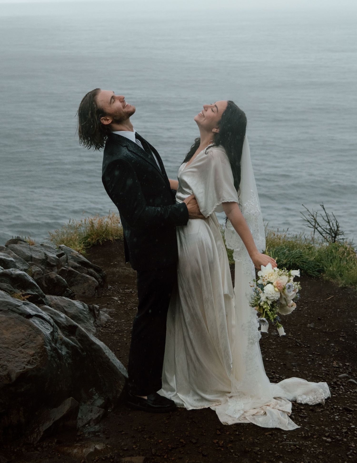 A wedding couple shares a joyful moment on a clifftop overlooking a misty ocean. Calgary Wedding Photographer