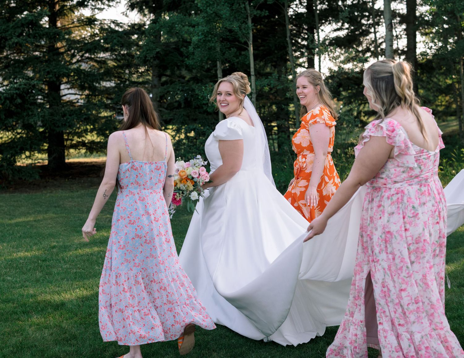 Group in floral pink dresses and white gowns walk through a lush green garden during a summer celebration.