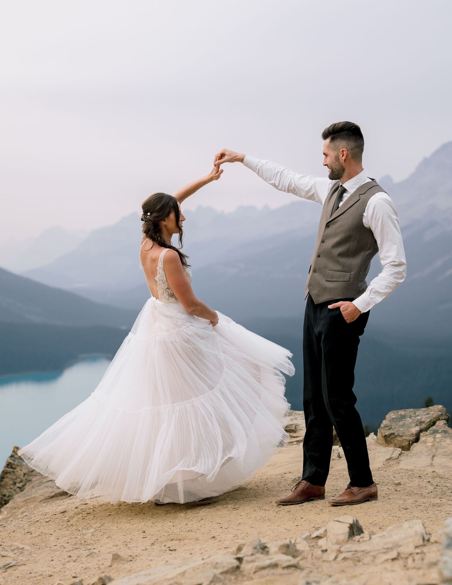 A romantic wedding couple poses dramatically on a mountain cliff overlooking a misty lake and mountain landscape.