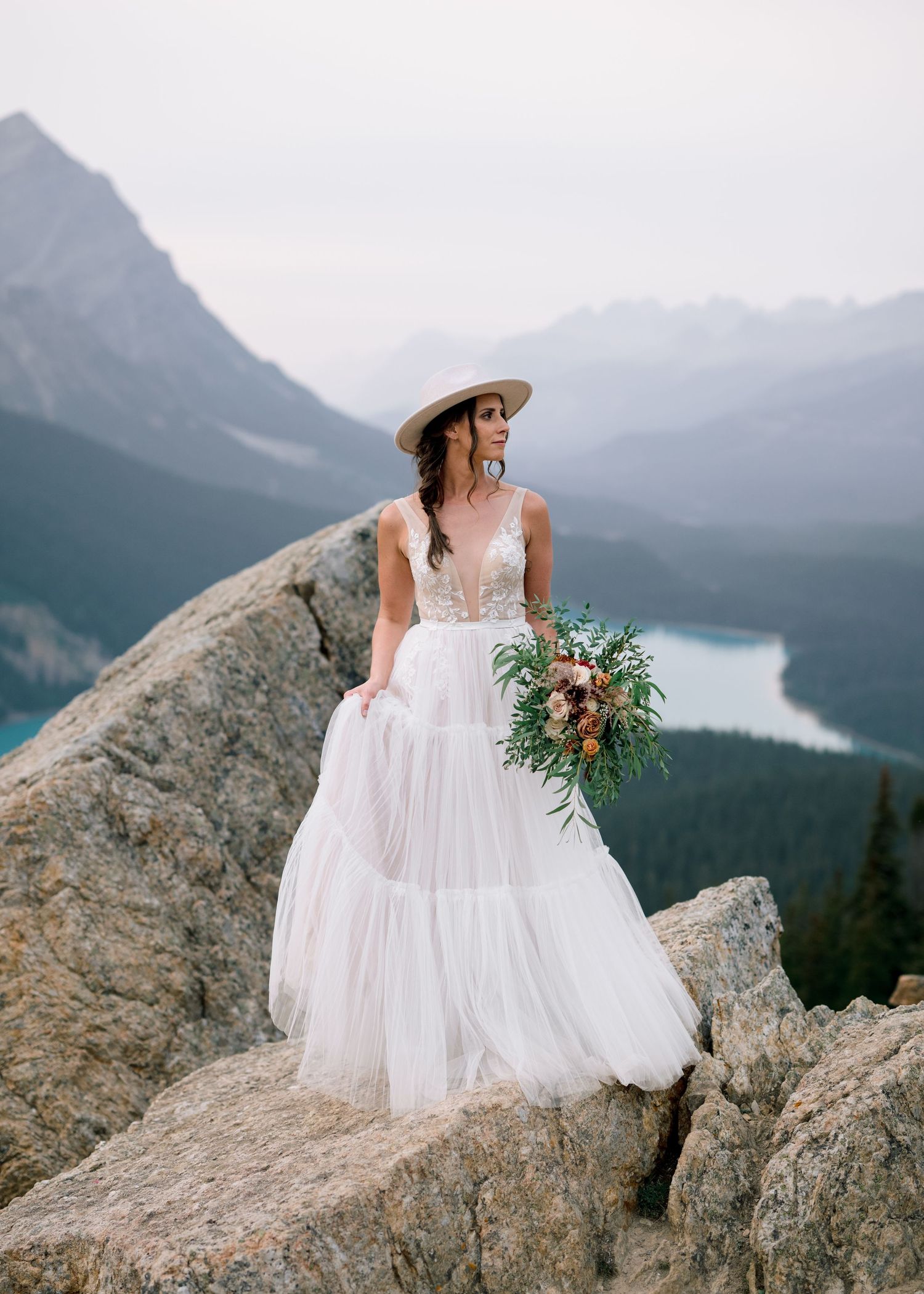A bride in a flowing white dress poses on mountain cliffs overlooking a turquoise lake and mountain vistas.