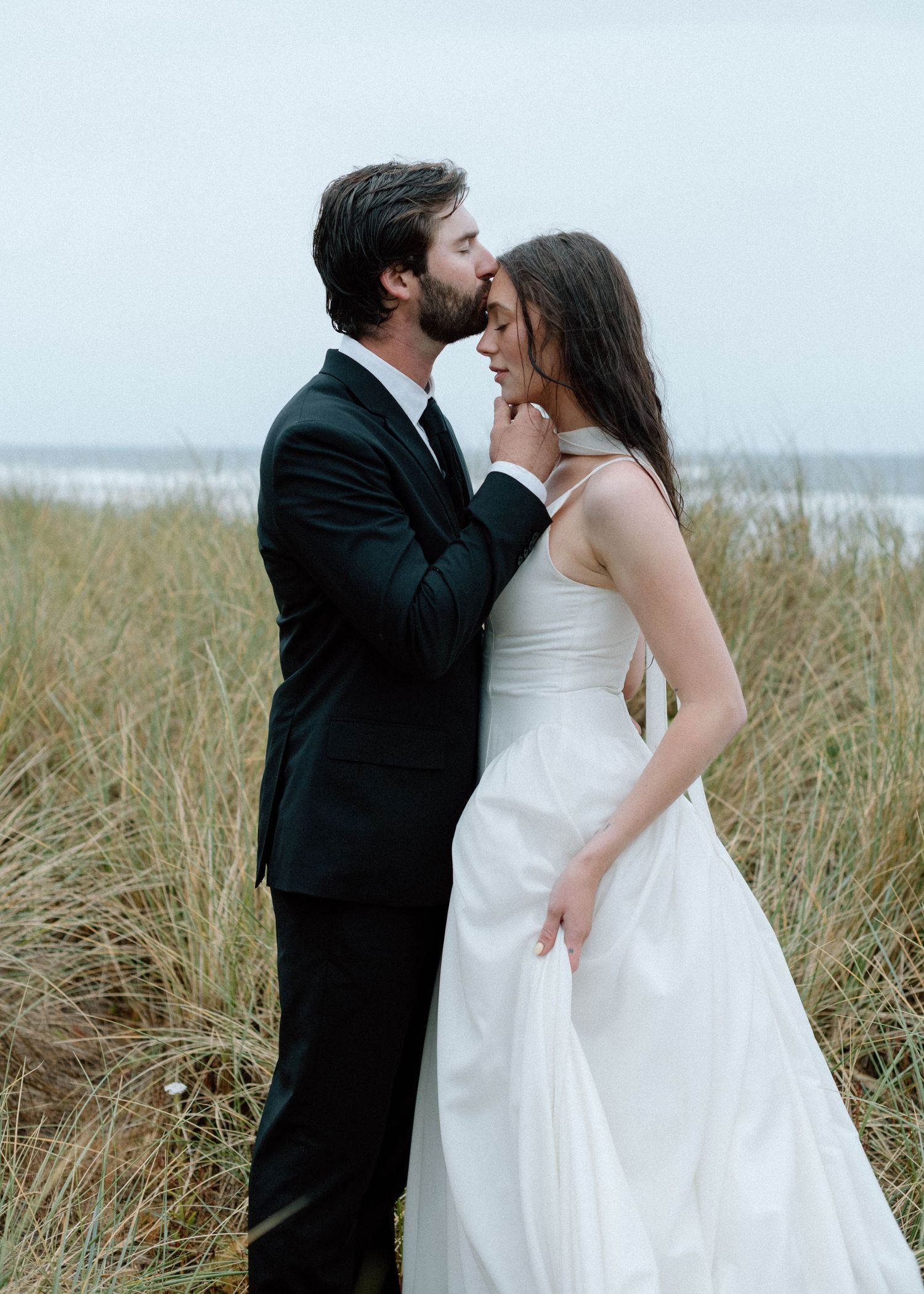 Newlywed couple embracing on a beach with tall grass in the foreground and ocean waves in the background.