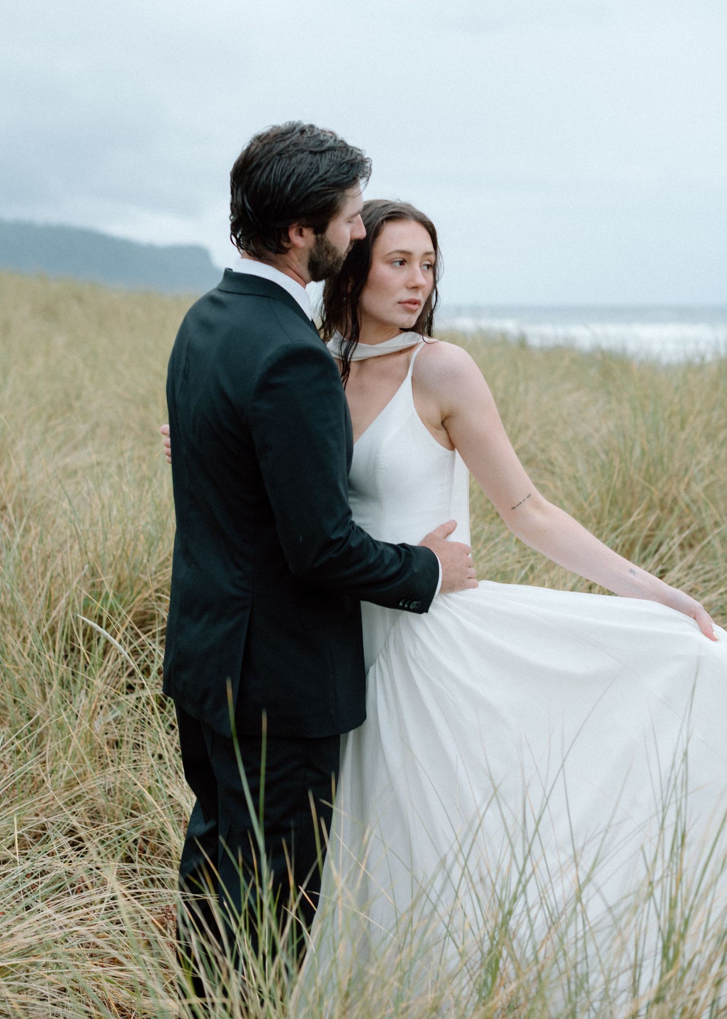Couple stands together among windswept beach grass on a cloudy coastal day.