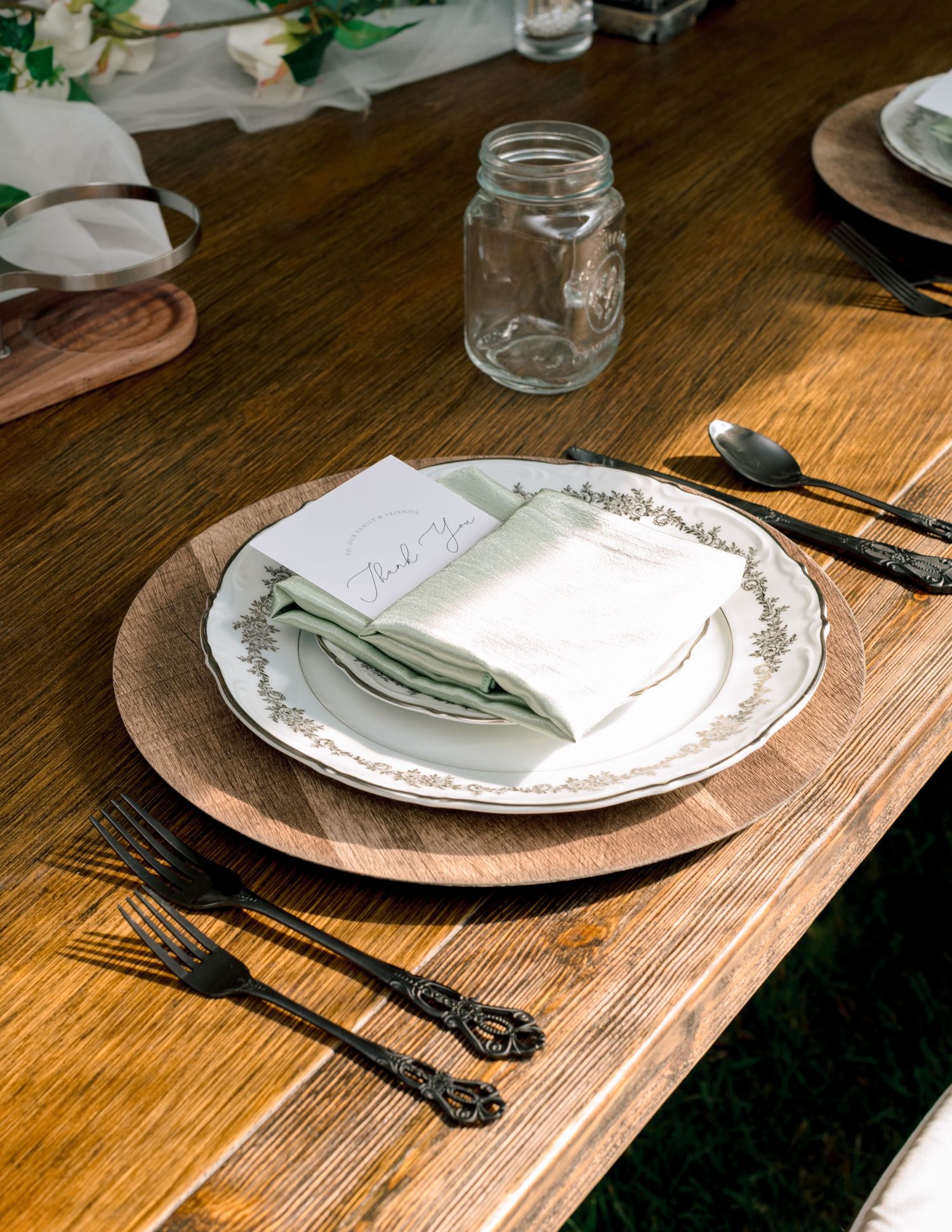 A rustic table setting with white plates on wooden chargers and sage green napkins in warm sunlight.