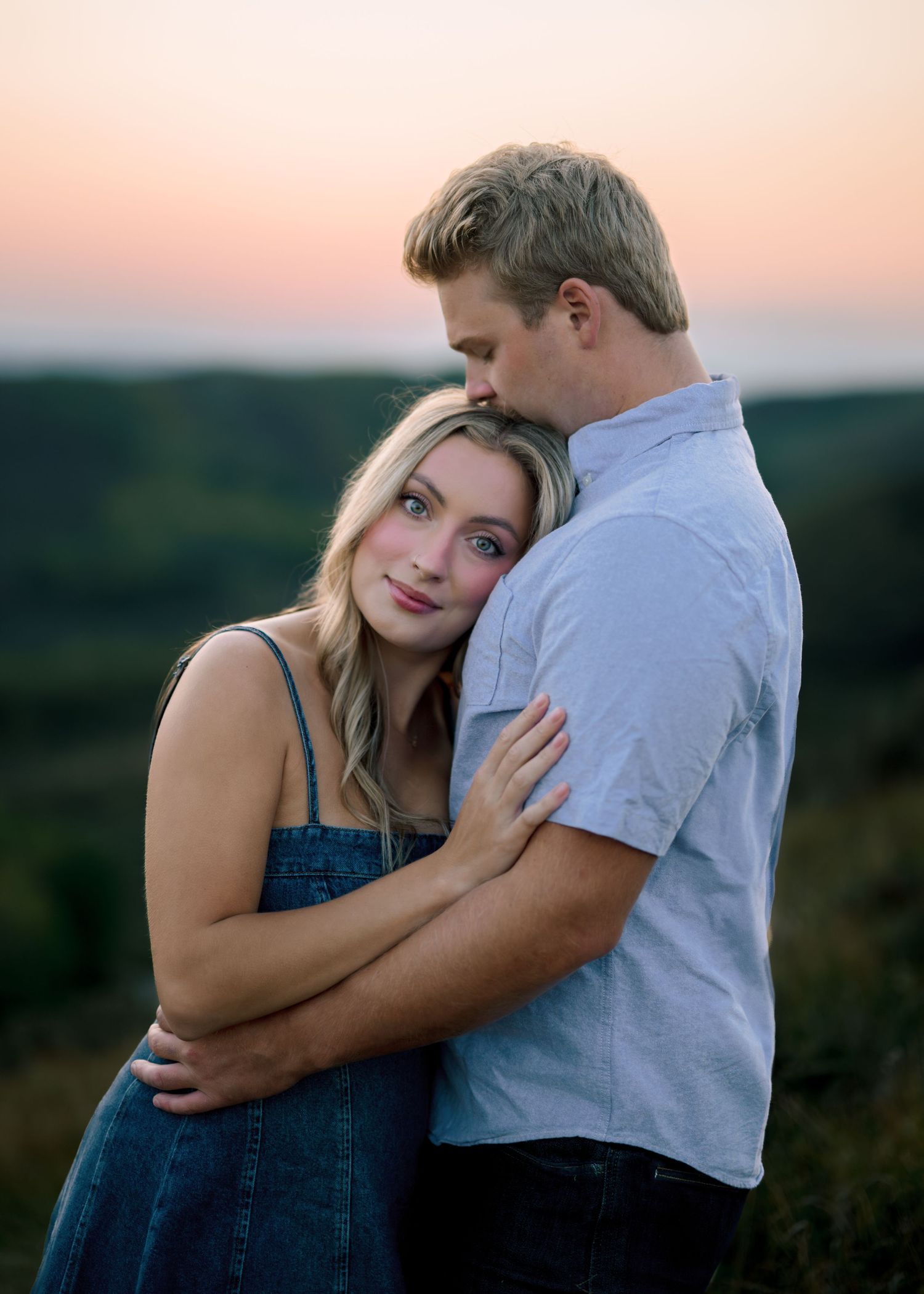 A couple in romantic embrace at sunset, with her in denim overalls and him in a light colored shirt against a dusky sky.