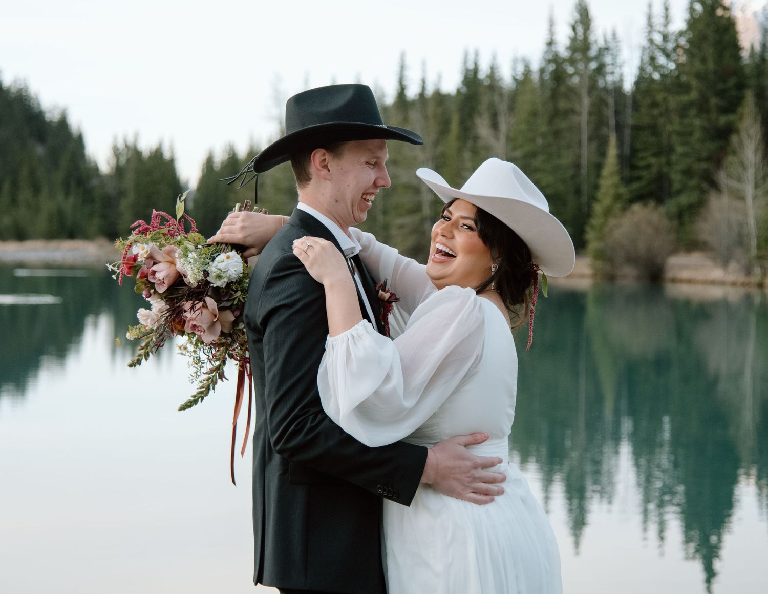 A romantic wedding couple embraces by a serene mountain lake with evergreen trees in the background. Banff and Calgary Wedding Photographer