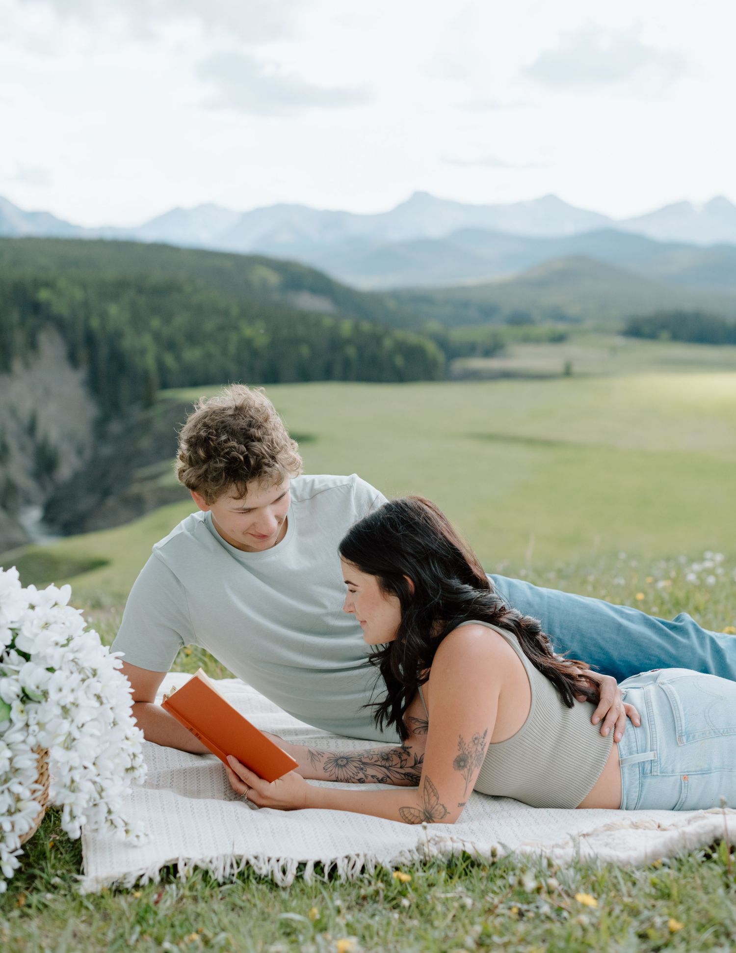 A couple lying in a mountain meadow reading together surrounded by wildflowers. Calgary Alberta Engagement Photographer.