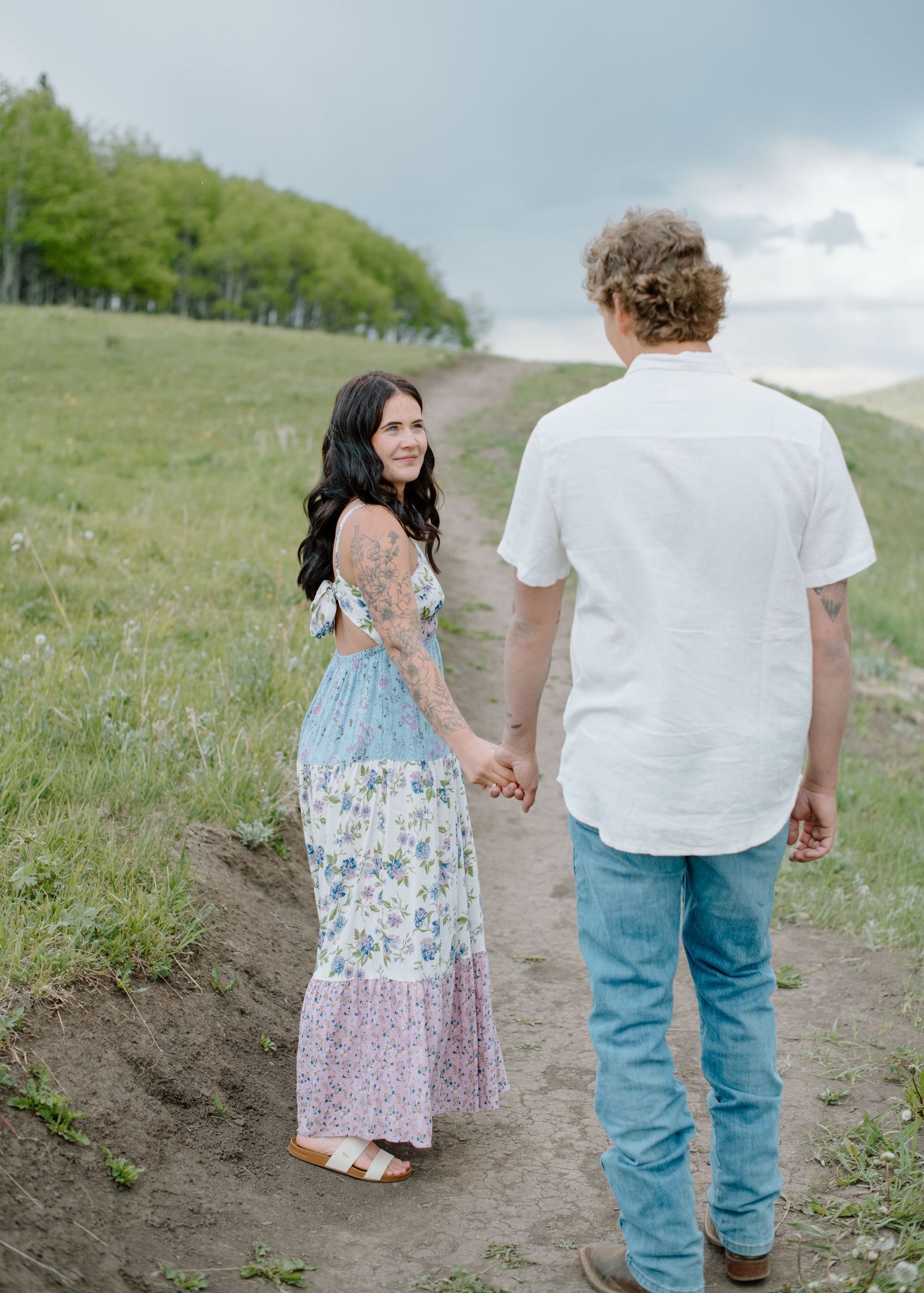 Couple walking hand in hand on dirt path through green rolling hills. Calgary Alberta Engagement Photographer.