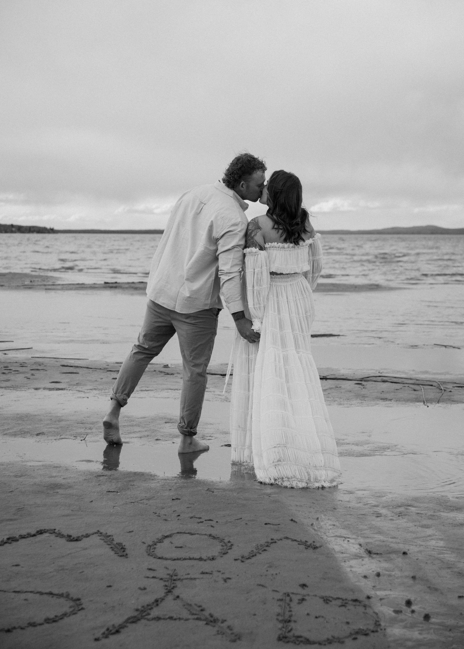 A black and white photo of a couple embracing on a beach with writing in sand. Calgary Alberta Maternity Photographer.