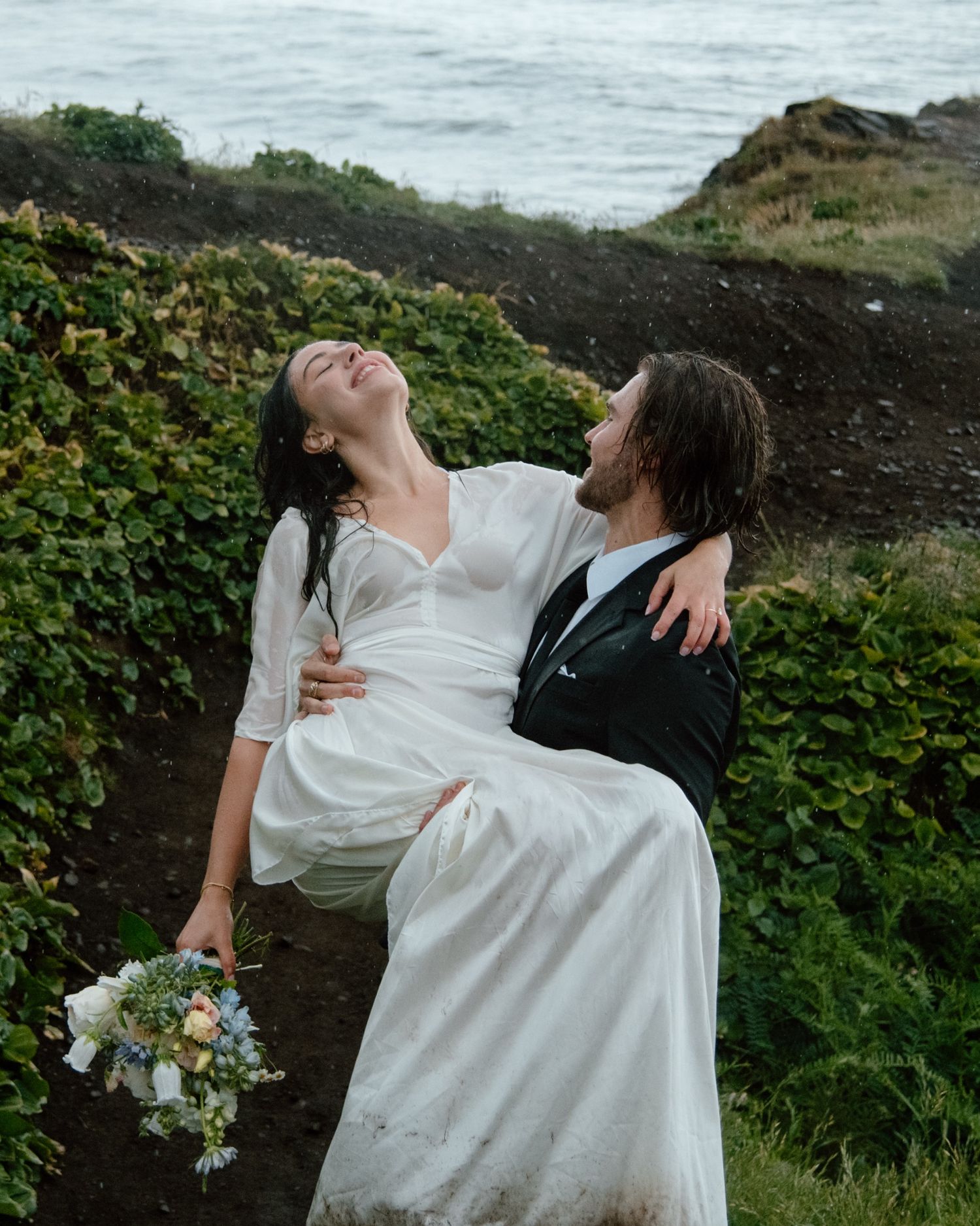 A couple embraces dramatically against a coastal clifftop backdrop with greenery and ocean views. Calgary Wedding Photographer