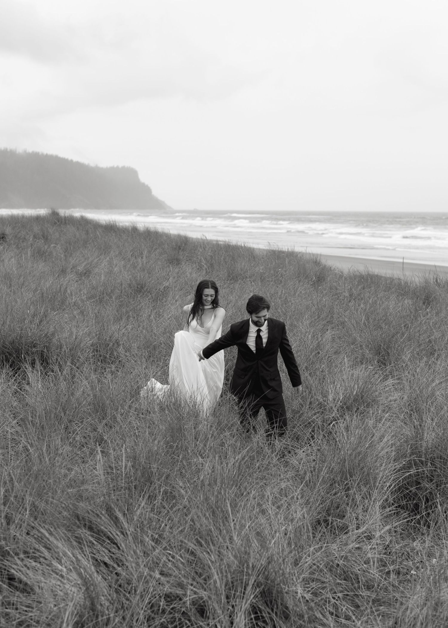 A couple runs playfully through tall coastal grass on a cloudy beach day during their wedding celebration.