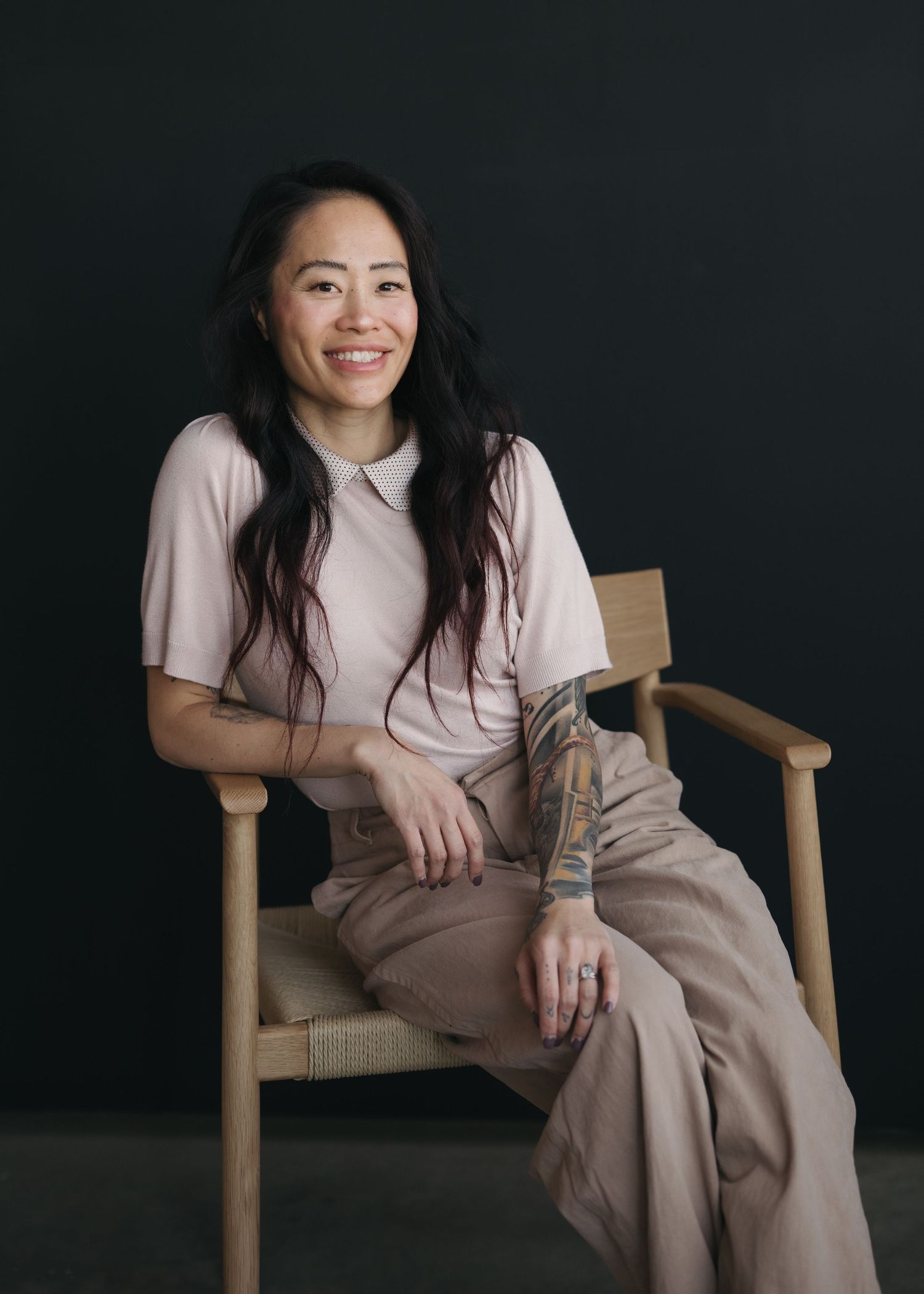 Person wearing a pink top and beige pants sits casually in a wooden chair against a dark background.