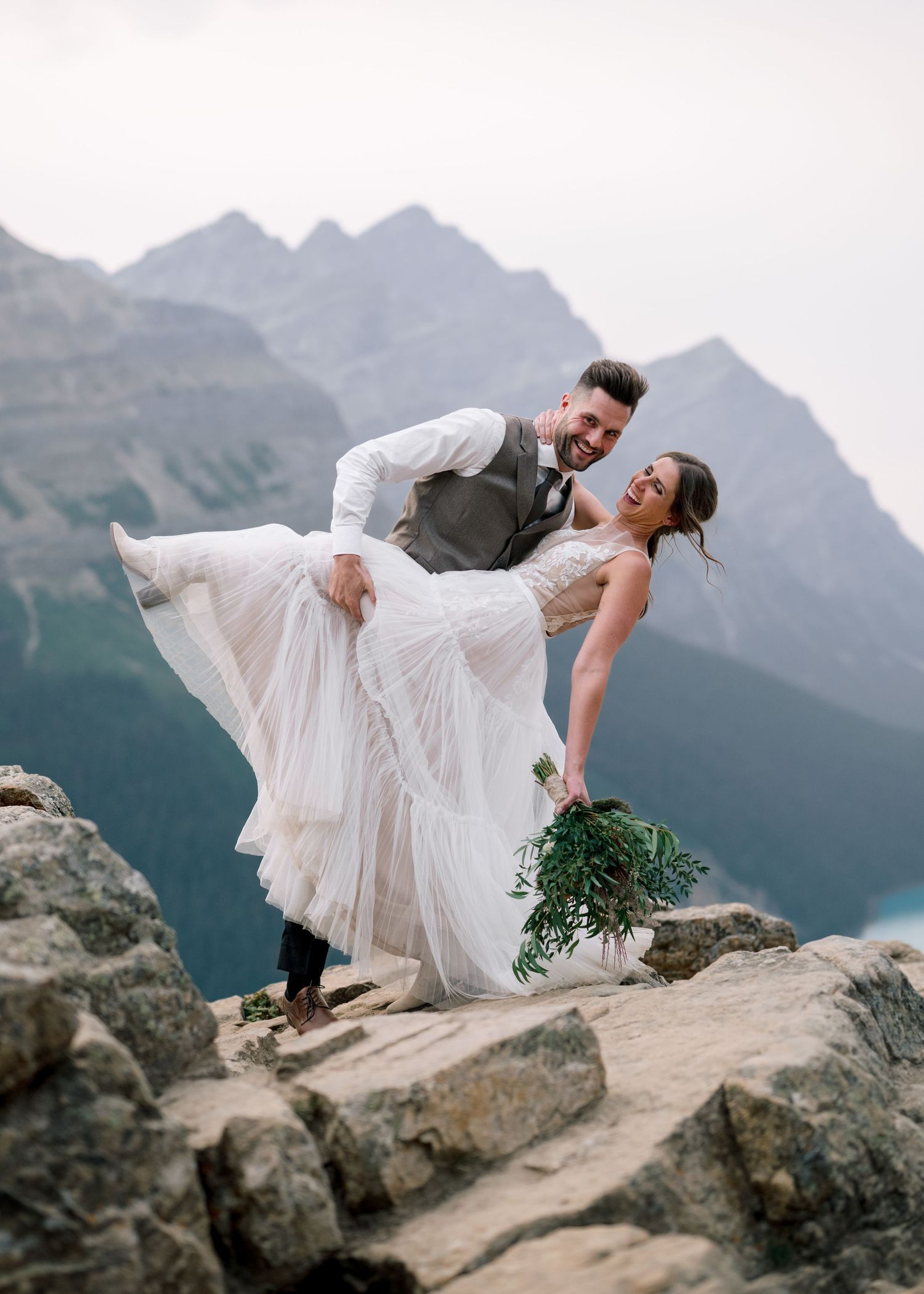 A couple dips dramatically on a mountain cliff edge with scenic mountain peaks and lake views in the background.