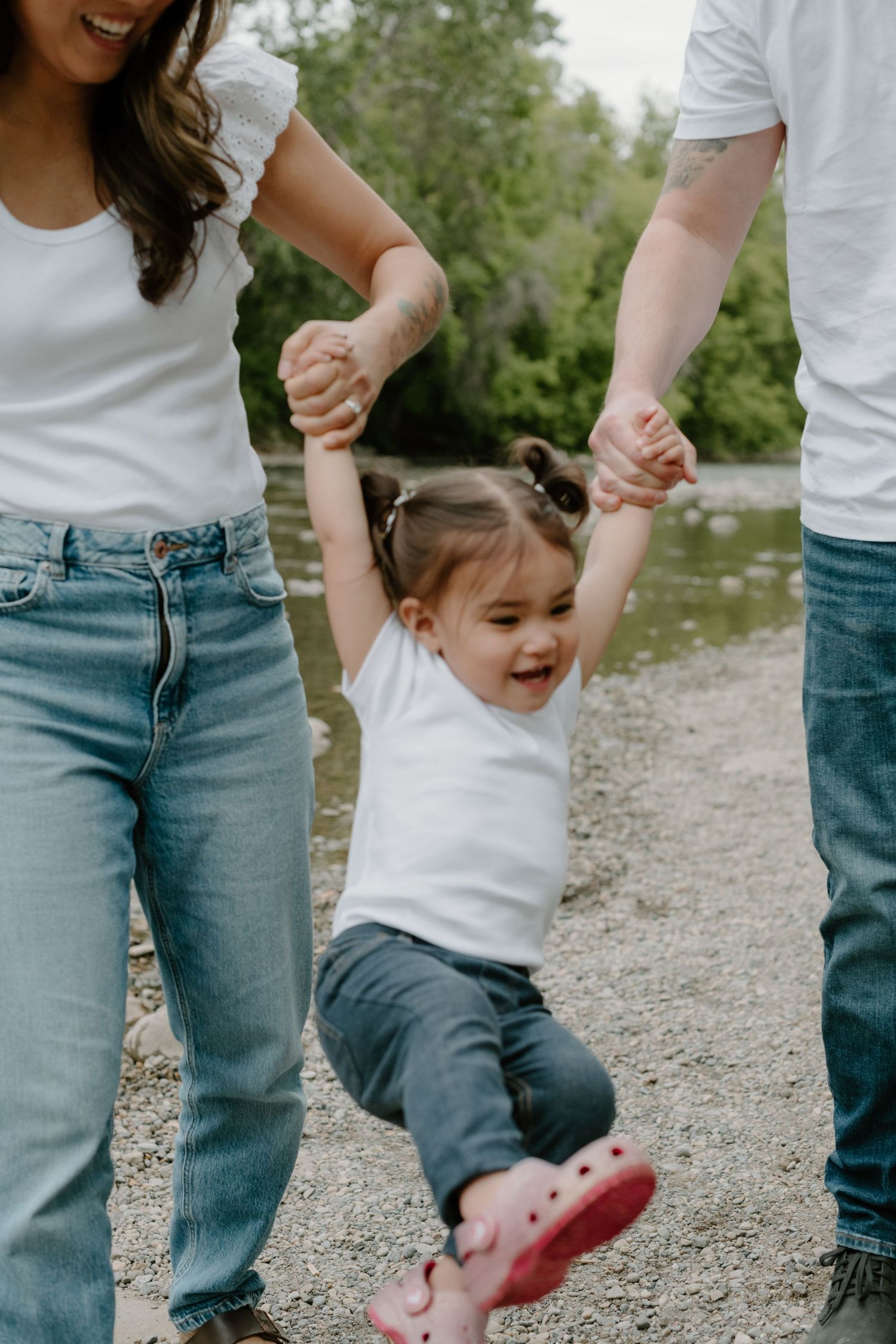 A young child swings joyfully between two adults on a riverside path. Calgary Alberta Family and Maternity Photographer.