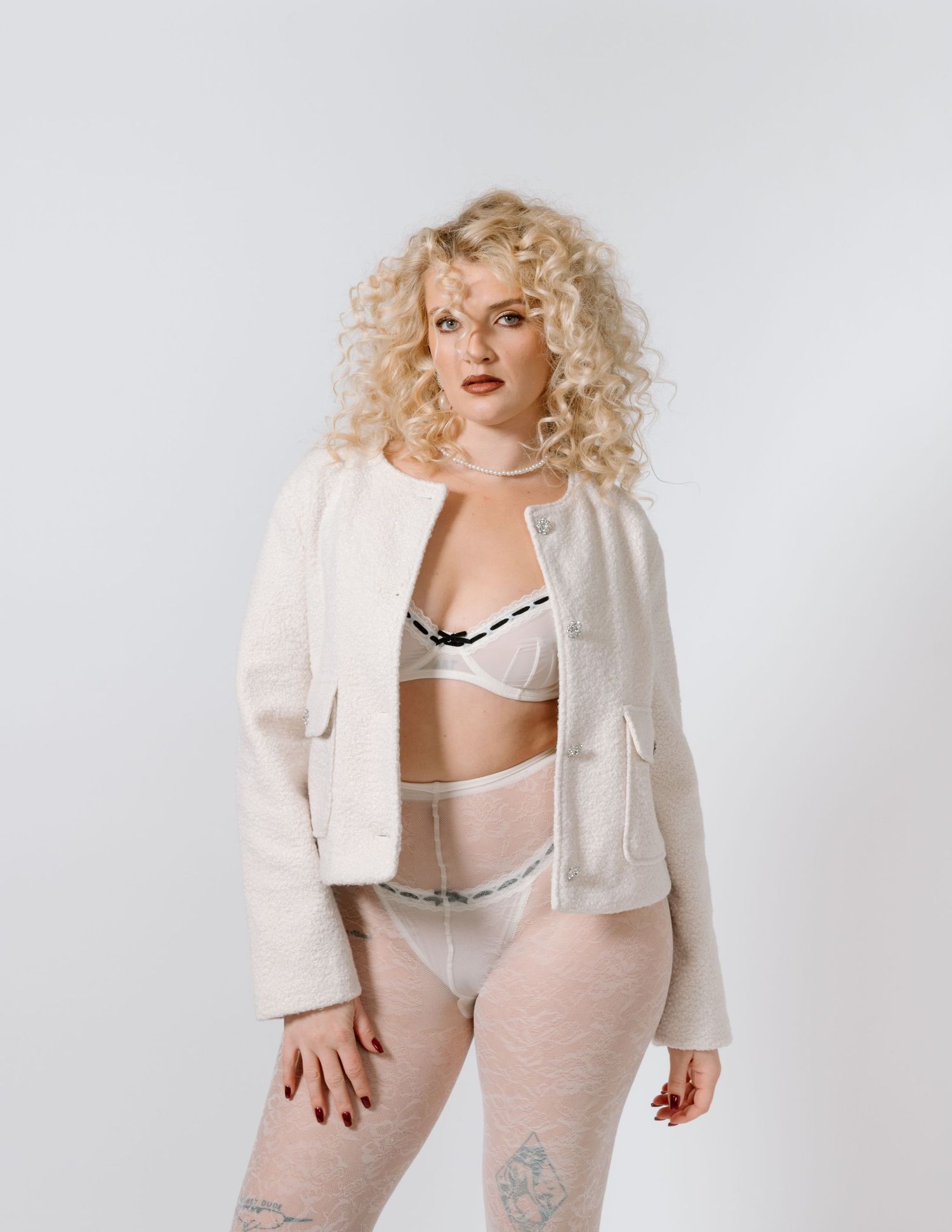 Model in white blazer and lingerie poses against neutral backdrop with voluminous curly hair. Calgary Alberta Bridal Boudoir Photographer.