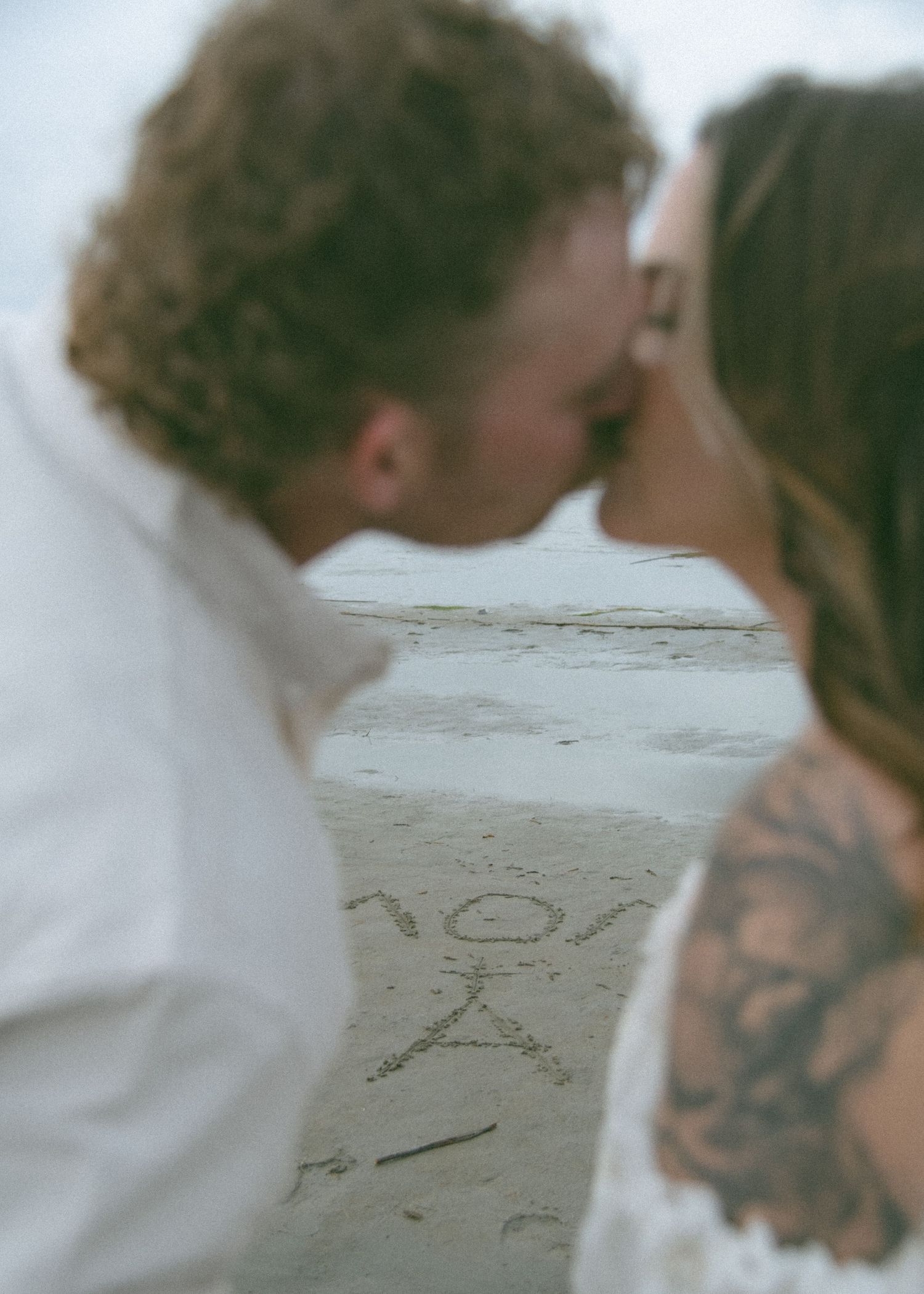 A romantic couple shares a kiss on a beach with text written in the sand. Calgary Alberta Maternity Photographer.